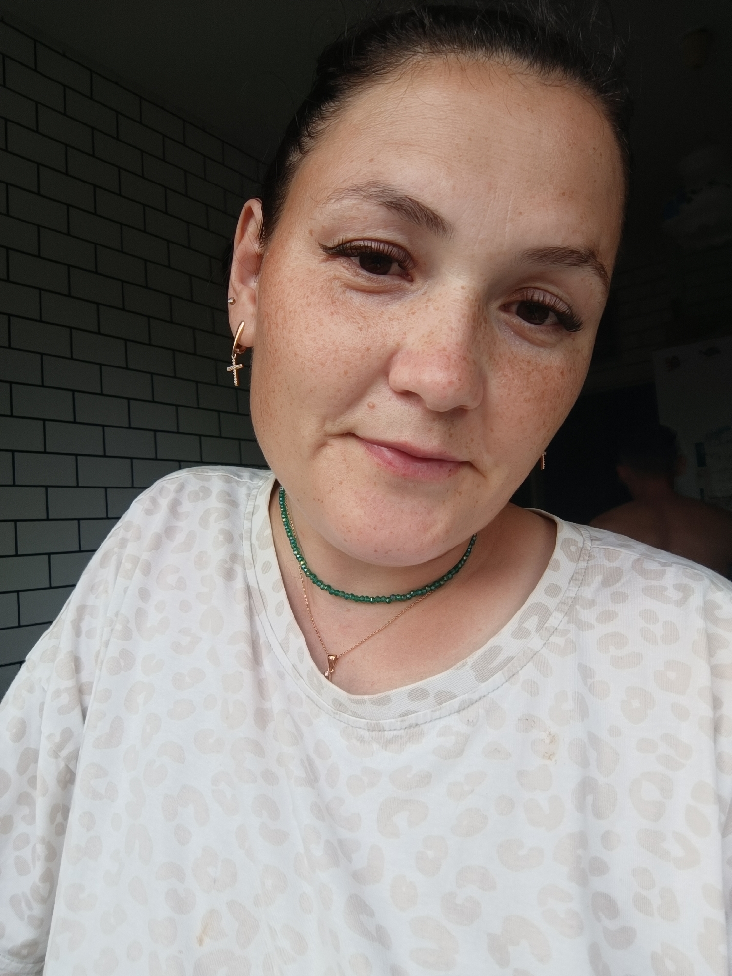 A woman with freckles is looking forward, her head slightly tilted. She has dark hair pulled back. She is wearing a white t-shirt with a leopard print pattern, a green beaded necklace, and a gold cross pendant. She has a gold hoop earring with a cross charm in her left ear. She is indoors, with a dark, brick-patterned wall behind her on the left and a darker, blurred background on the right. There is a person's bare back visible in the blurred background on the right. The scene appears to be in a domestic setting, possibly a kitchen or living area. The image does not contain any specific indicators of location in Chaikovsky, Russia.