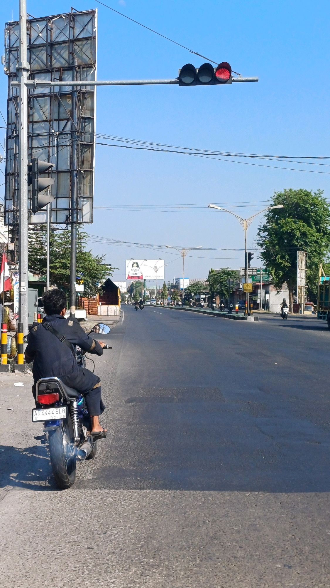 A motorcycle rider, wearing a dark blue jacket and pants, is positioned in the foreground, facing away from the camera and looking towards a road. The rider is on a dark blue motorcycle with a license plate that reads "AD 4444 ELO". The scene is a street in Ngadirejo, Indonesia. In the background, traffic lights show a red signal, indicating that vehicles should stop. The sky is clear and blue. The road is paved and appears to be a busy thoroughfare with other vehicles and motorcycles visible in the distance. There are buildings and trees lining the sides of the road. A large billboard stands on the left side of the image, and streetlights are visible along the road. The overall impression is a typical urban street scene on a sunny day.