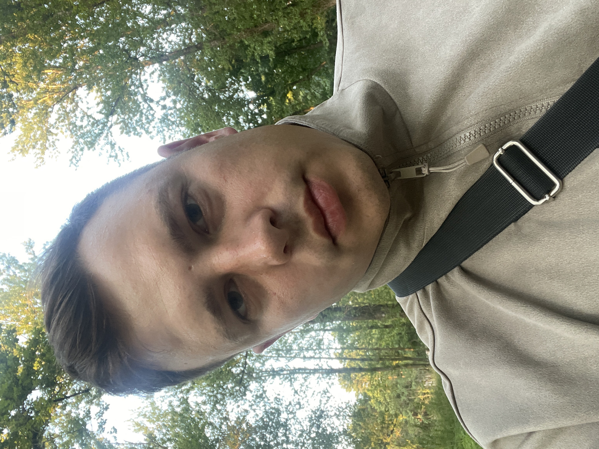 The media file shows a close-up, vertically oriented selfie of a person from the chest up, outdoors. The subject appears to be a young man with short, dark hair and fair skin. He is wearing a beige-colored zippered jacket with a black strap across it, possibly from a bag. The background consists of trees with green foliage, suggesting a natural, possibly wooded, setting. The lighting indicates it is daytime, with sunlight filtering through the leaves. The overall mood of the image is casual and relaxed. No text is visible in the media file.
