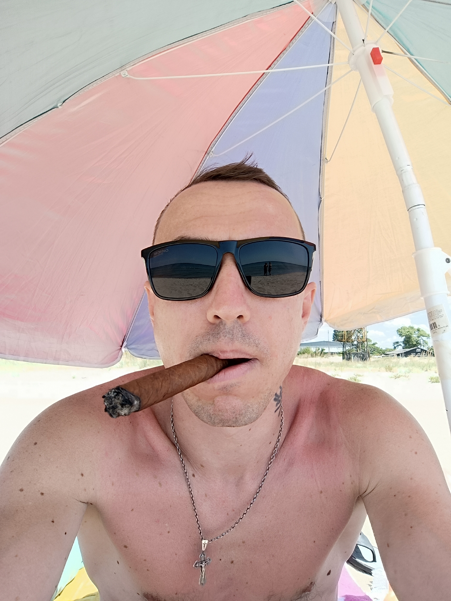 A shirtless man wearing sunglasses and a silver chain with a cross pendant is shown in a close-up, selfie-style shot. He is holding a lit cigar in his mouth. The man is positioned under a multicolored beach umbrella, with segments of pink, light blue, and yellow fabric visible. Behind him, a sandy beach and a body of water are visible. In the background, there are trees and some buildings, including a structure that appears to be a lifeguard tower. The man's skin shows signs of sunburn on his shoulder. The image appears to be taken in Zatoka, Ukraine, a seaside resort town.