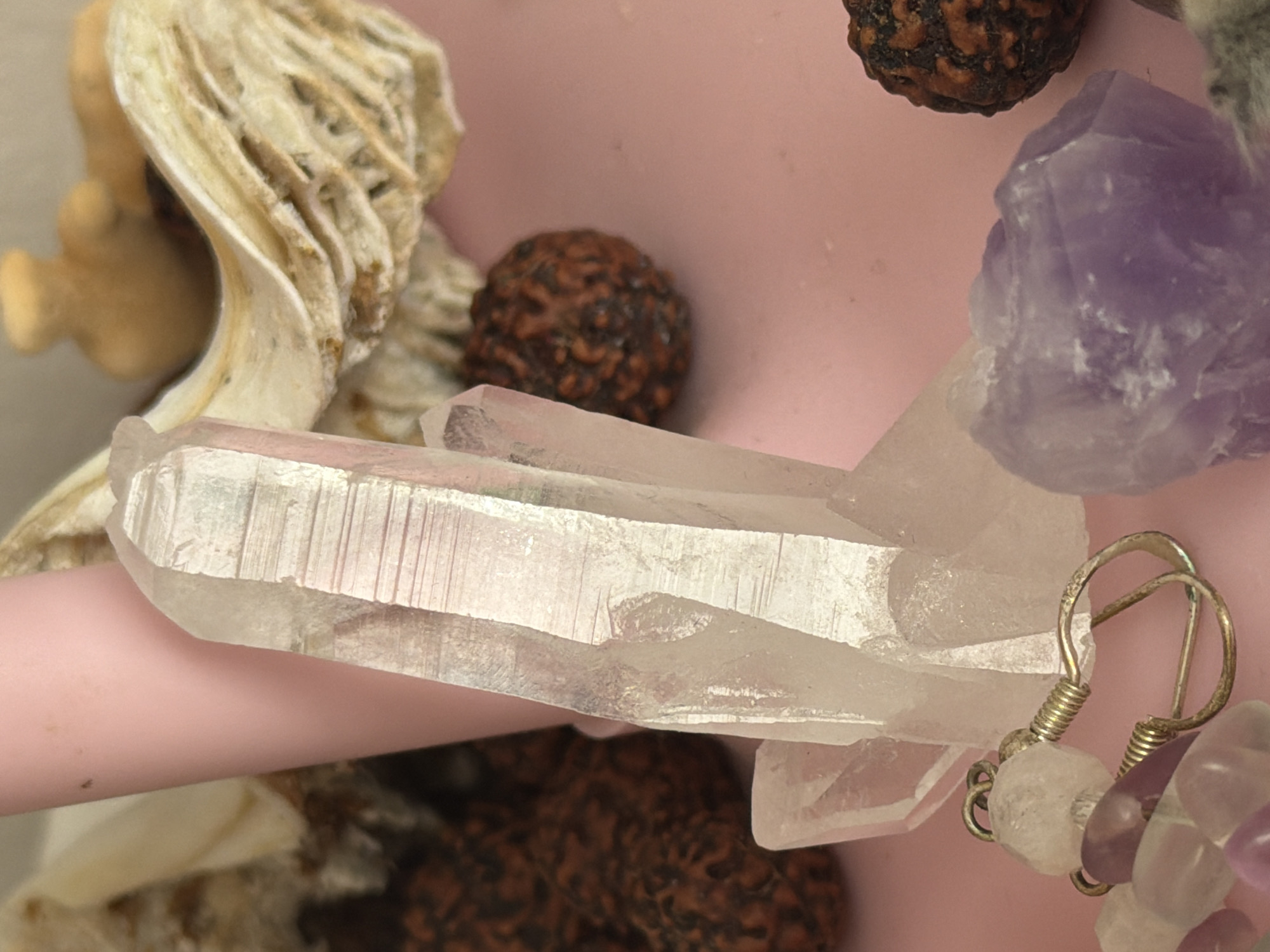 The image features a collection of objects, including a white, crystalline quartz crystal cluster, a piece of amethyst, brown rudraksha beads, a piece of what appears to be a shell, and a silver earring with beads. These items are placed on top of a pink surface. No information within the image indicates that it was taken in the city of Saint Petersburg, Russia.