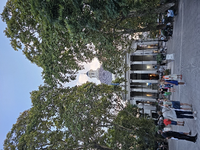 The image depicts the Basílica Nuestra Señora del Pilar, a neoclassical church located in the Recoleta neighborhood of Buenos Aires, Argentina, at dusk. The basilica stands centrally, framed by two large, leafy trees on either side which partially obscure the upper corners of the structure and sky.
The basilica features a prominent white facade with a portico supported by tall, fluted columns and a large entrance behind them. Above the main structure, a circular dome with a patterned blue and white mosaic or tiled surface is visible, topped by a white lantern structure culminating in a cross. An Argentinian flag, light blue and white, hangs from a flagpole attached to the upper left portion of the basilica's facade. Artificial lighting illuminates the building, enhancing its features against the fading natural light of the twilight sky, which is a clear, light blue.
In the foreground and mid-ground, a wide, patterned paved plaza extends, featuring light and dark grey pavers. Approximately twenty to thirty individuals, predominantly adults in casual attire, are present on the plaza. Several small groups of people are standing and conversing in the mid-ground, facing various directions. One individual in a black shirt and trousers stands with their back to the viewer, facing a small group of people. Other individuals are scattered closer to the basilica's entrance or seated on benches or the ground near the base of the trees on the far right. The overall scene suggests a public gathering or leisure activity in a well-maintained urban environment.
