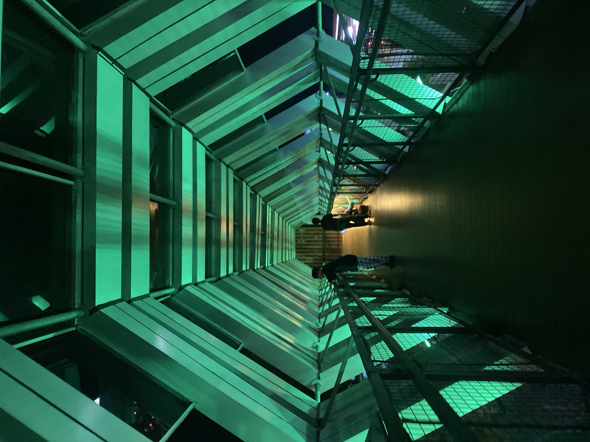 This image depicts the enclosed pedestrian bridge of the Banjarbaru Skatepark in Banjarbaru, Indonesia, illuminated by vibrant green lights. The bridge has a hexagonal, tunnel-like structure with angled panels on the ceiling and sides, creating a geometric and futuristic aesthetic. A metal railing runs along both sides of the walkway, featuring a mesh design that allows the green light to cast diamond-shaped patterns on the walkway and the exterior. Two individuals are visible on the bridge. The first, on the left, is a person wearing a dark top and checkered pants, leaning on the railing and looking out. Further down the bridge, another person, silhouetted against the light, is walking away from the camera. The walkway appears to be made of dark wood planks. The overall ambiance is moody and modern, highlighting the architectural design of the bridge with its dramatic lighting.