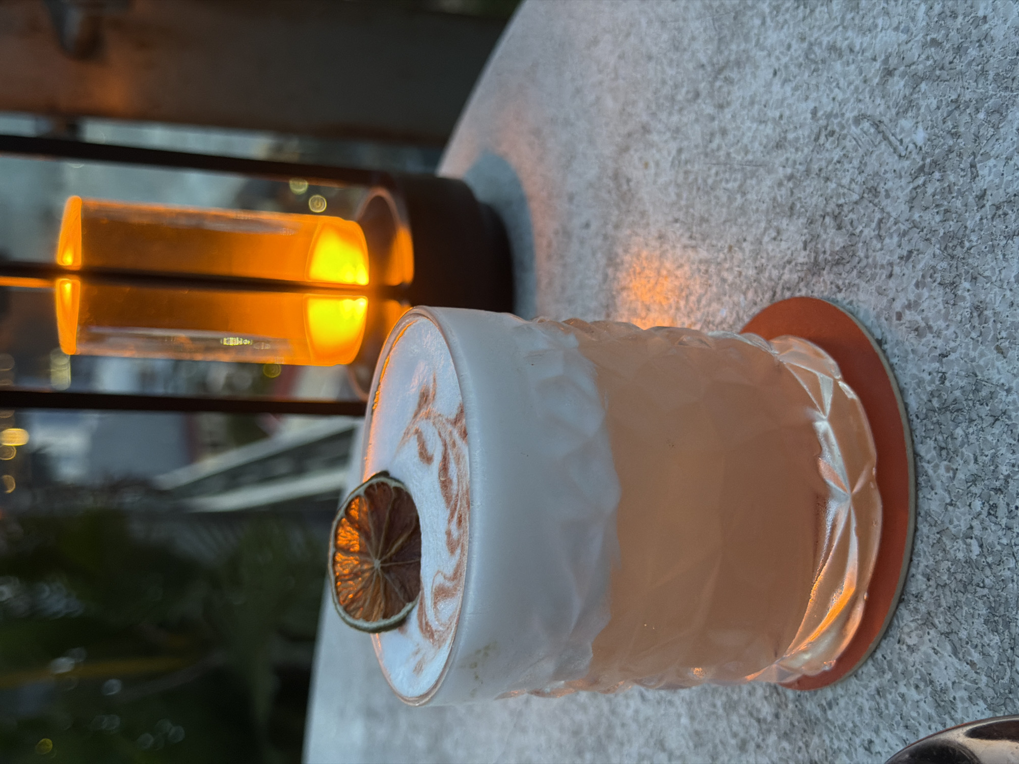 A close-up shot features a cocktail with a foamy top, garnished with a dried lime wheel, resting on a textured grey surface. The drink is in a clear, faceted glass with a light orange hue. Behind the drink, an illuminated lantern with an orange glow is visible. The background is blurred, showing greenery and distant lights, suggesting an outdoor setting at dusk or night, possibly in Đà Nẵng, Vietnam, given the presence of tropical vegetation and city lights. The overall atmosphere is cozy and inviting.