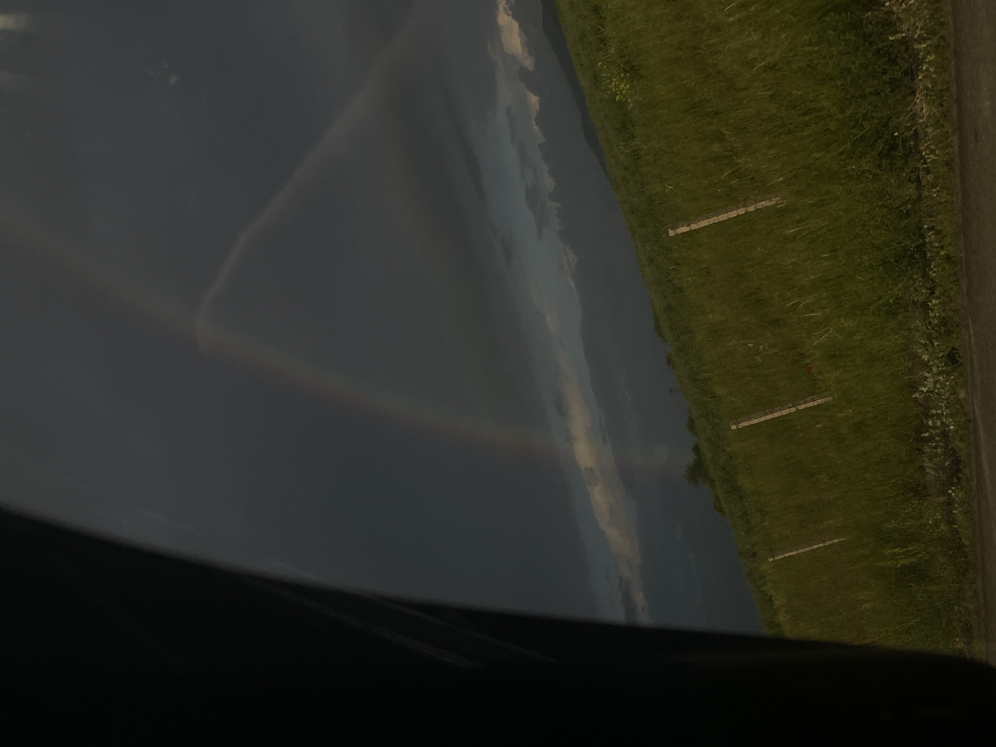The image captures a rural landscape in Dragichevo, Bulgaria, observed from a vehicle, with a dark, vertical structural element partially obscuring the far left. The sky is predominantly overcast with dark grey clouds, beneath which a luminous partial rainbow arc is visible in the upper-middle. A vibrant green hillside, densely covered with tall grass and various foliage, slopes from the center-right towards the upper-right. Several light-colored wooden fence posts are irregularly spaced along this grassy incline. A narrow, dark strip, consistent with a road surface, is visible at the bottom edge of the frame. The overall lighting is subdued, characteristic of an overcast or post-rain environment. No individuals or animals are present within the scene, and no explicit actions are depicted.