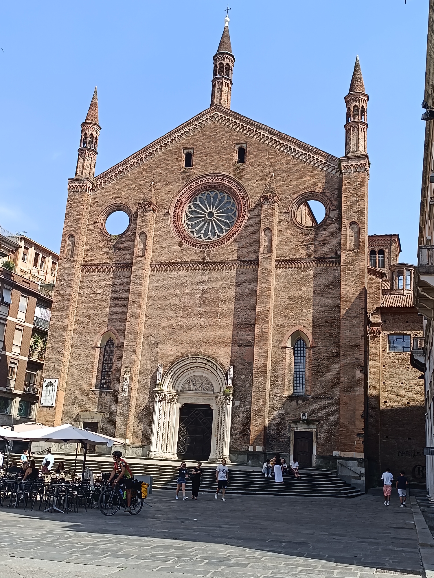 The image shows the Basilica of Sant'Antonino in Piacenza, Italy. The basilica, built of red brick, is viewed from its front facade. It has a central rose window and two narrower circular windows on either side. Twin towers flank the central peak of the roof. A grand entrance with a carved tympanum is at the base of the basilica. Wide steps lead up to the entrance.
In the foreground, a stone-paved square is populated with people. Some are sitting at tables under a white umbrella, others are walking across the square, and a cyclist with panniers rides by. Buildings adjacent to the basilica are visible on either side. The sky is clear and blue.