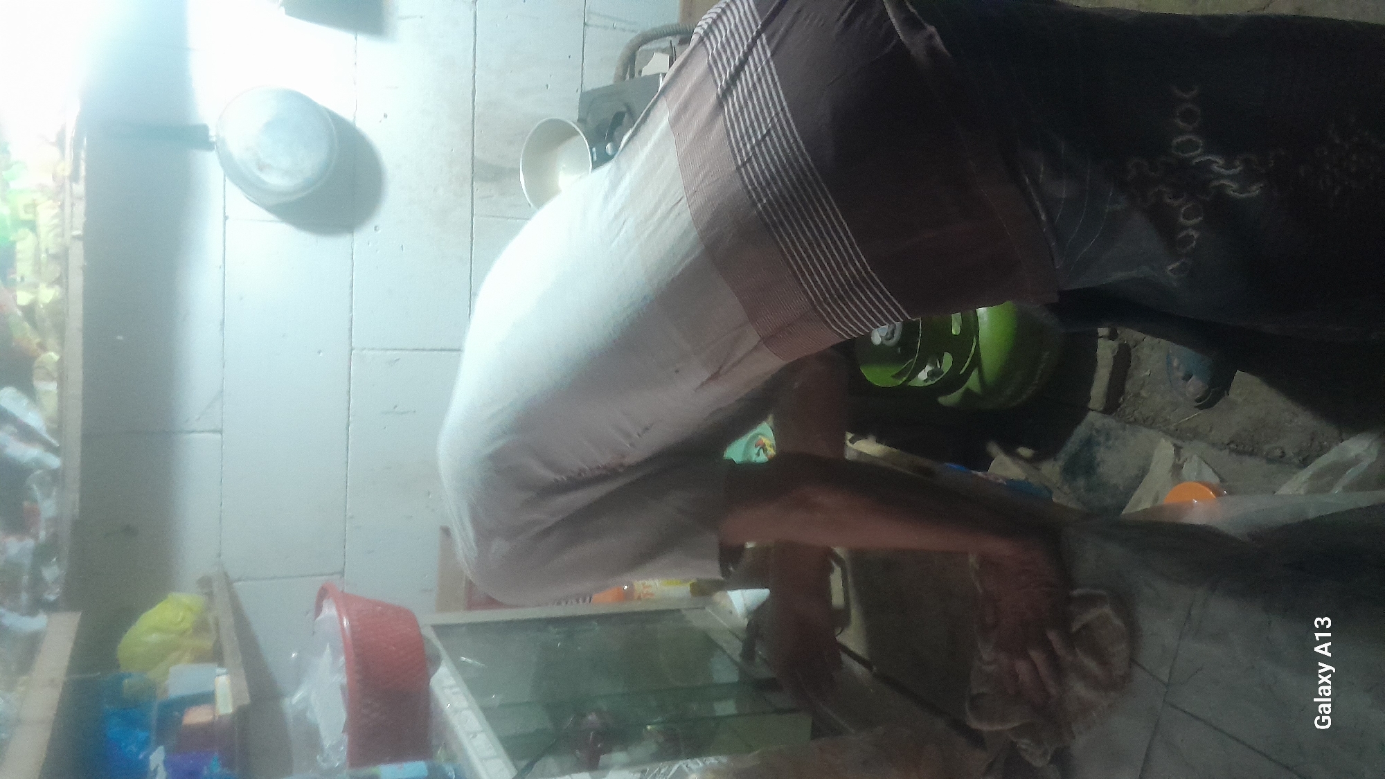 A person is bent over in a dimly lit room, appearing to be in the process of cooking or cleaning. They are wearing a light-colored shirt with a dark patterned lower garment. A green gas cylinder is visible behind the person, along with various household items and shelves in the background. The scene suggests a domestic environment, potentially a kitchen or utility area. The "Galaxy A13" watermark indicates the image was captured with a Samsung Galaxy A13 smartphone. The specific location is stated as Hadipolo, Indonesia, though the visual evidence does not provide further details to corroborate this.