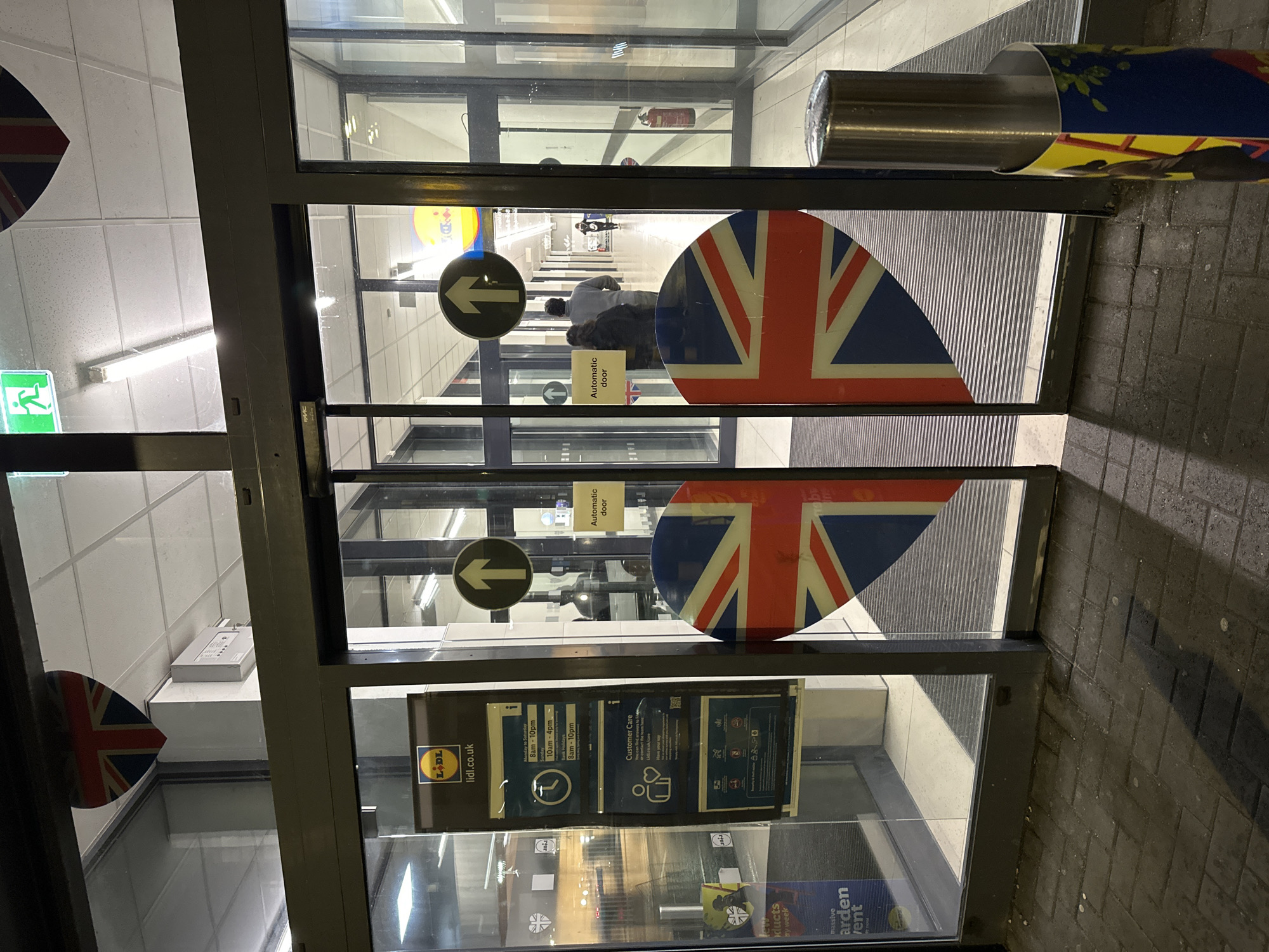 The image depicts the exterior of a Lidl supermarket entrance in London, United Kingdom, under low-light conditions, suggesting evening or night. The entrance features dark-framed automatic glass sliding doors with fixed glass panels on either side.
On the lower half of the two central sliding doors, two large heart-shaped decals displaying the Union Jack flag are prominently affixed. Smaller, similar heart-shaped Union Jack decals are visible higher up, above the main door frame, on the left and right. Each central sliding door also bears a circular black sign with a white upward-pointing arrow, indicating entry, and a rectangular white sticker stating "Automatic door."
To the left of the main entrance, on a fixed glass panel, a black vertical sign holder displays the yellow Lidl logo with "lidl.co.uk," followed by detailed store opening hours (e.g., "Monday to Saturday 8am - 10pm," "Sunday 10am - 4pm") and "Customer Care" information, including a QR code.
Through the transparent doors, a brightly illuminated interior corridor or aisle of the supermarket is visible, featuring a white tiled ceiling with fluorescent lighting. Two individuals are seen inside, walking away from the entrance: one wearing a light-colored top, with another person in darker attire partially obscured behind them. A third person is discernible further down the corridor. A red fire extinguisher is mounted on the wall to the right within the interior corridor. An illuminated green exit sign is visible above the interior threshold.
The ground immediately outside the entrance consists of dark grey block paving. To the right of the entrance, a silver cylindrical bollard is present, partially covered with a colorful graphic wrap featuring yellow, blue, and green elements, possibly advertising.