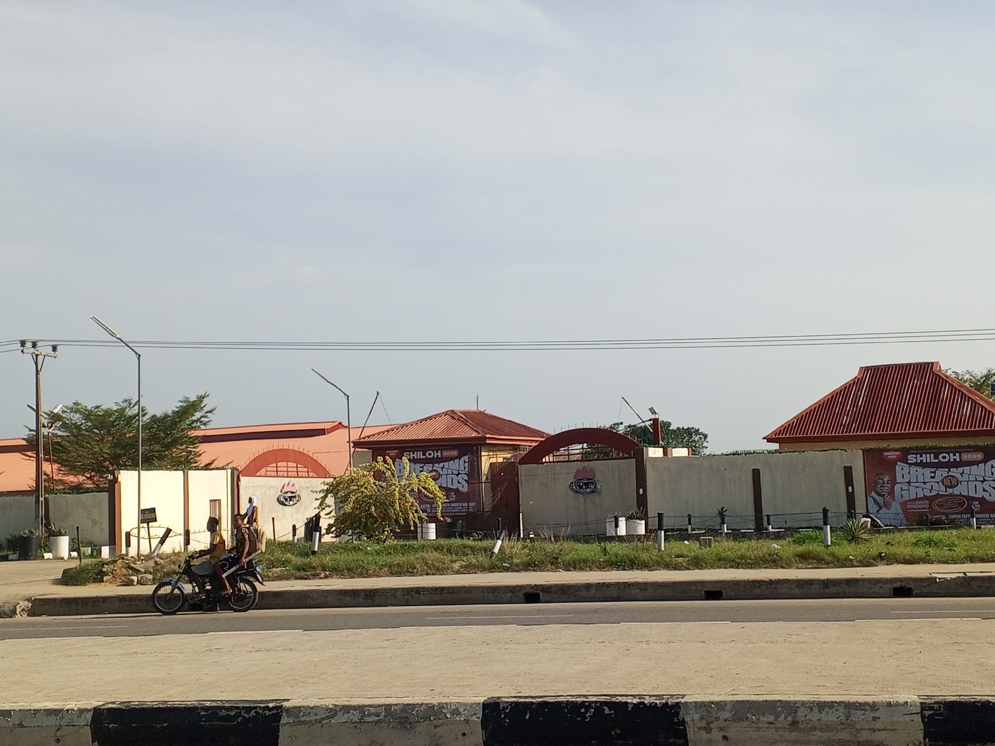 This daytime image, set in Ajara, Nigeria, presents a view across a multi-lane road towards buildings set back from the street. In the foreground, a concrete curb painted with black and white stripes runs horizontally. On the road, slightly to the left, a motorcycle with three occupants – two adults and a child – is stopped or moving slowly.
Beyond the road, a grassy median separates the thoroughfare from a series of structures. Several light poles and overhead power lines are visible against the sky. The background features multiple buildings with reddish-brown, ridged roofs. These buildings are largely obscured by a light-colored concrete wall or fence that runs along the property line. Green trees and bushes are scattered throughout the scene, adding natural elements.
Two prominent banners are affixed to the concrete wall. Both display "SHILOH 2023" and "BREAKING NEW GROUNDS." The banner on the right also features a portrait of "BISHOP DAVID OYEDEPO" and smaller text likely indicating "LIVING FAITH CHURCH WORLDWIDE." A white circular sign with an indiscernible logo (possibly "SWEET FRIES") is also visible on the wall to the left. The sky is clear with light cloud cover, suggesting bright daylight, likely late morning or early afternoon, with no strong shadows.