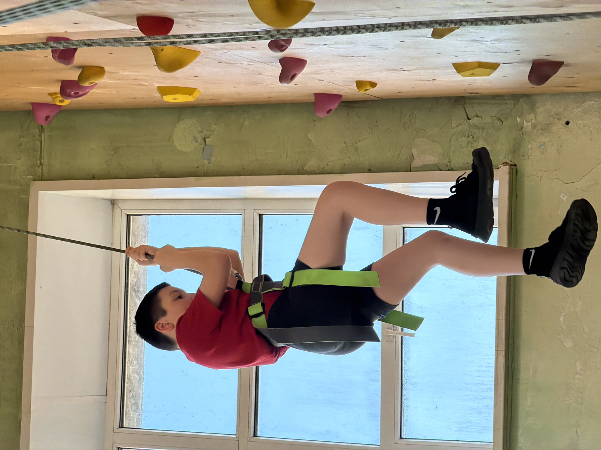 A young boy is captured suspended horizontally in an indoor climbing or training facility. He is dressed in a red t-shirt, black shorts, and black socks with black sneakers; a white Nike swoosh logo is visible on his left sock. A green and black climbing harness secures him, linking him to a dark-colored rope that he holds with both hands. His body is parallel to the floor, with his head turned slightly, showing a concentrated expression.
Above him, a light-colored wooden ceiling is studded with numerous climbing holds in vibrant red, yellow, and pink. Another rope is strung across these ceiling holds. The background features a distressed, pale green wall with visible cracks and flaking paint. A large window fills the lower portion of the frame, allowing bright natural light into the space and revealing a clear blue sky outside.
The activity appears to be an advanced climbing or aerial training exercise, demanding strength and focus. The setting suggests a dedicated indoor climbing gym or a specialized training room in Izhevsk, Russia, observed during daytime hours due to the natural light.