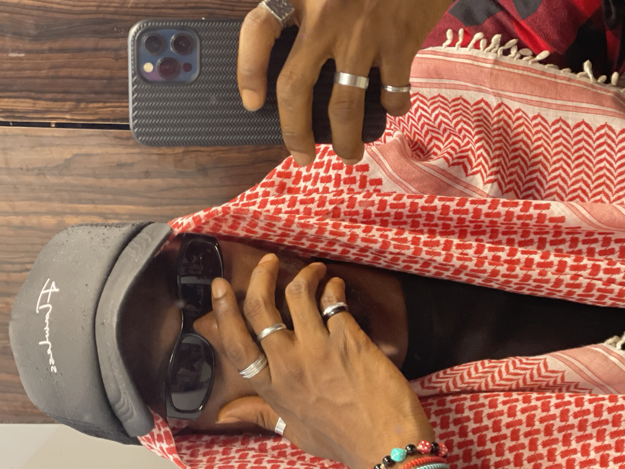 A close-up, waist-up shot shows a person holding a smartphone and taking a selfie. The person is wearing a black baseball cap with white lettering, black sunglasses, and a red and white patterned keffiyeh. The keffiyeh is draped around their neck and shoulders, partially covering a black t-shirt and a red and black plaid shirt. Multiple silver rings are visible on the person's fingers, and a beaded bracelet is on their left wrist. The background appears to be a wooden surface, possibly a wall or door, with a vertical seam running down the center. The lighting suggests an indoor environment. There is no discernible context to place this image in Abuja, Nigeria.
