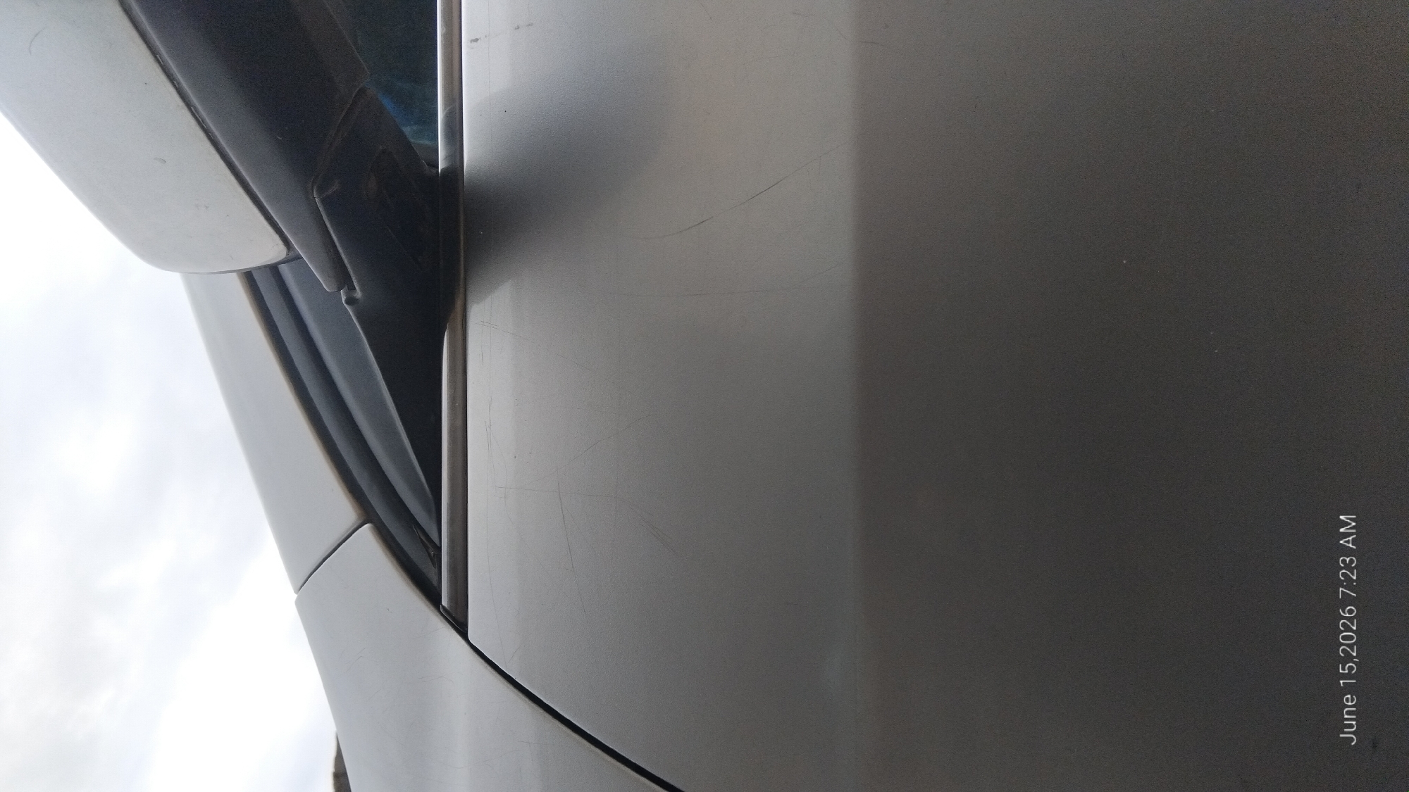 The image shows a close-up, low-angle shot of a silver car door and side mirror, with a cloudy sky in the background. The car's paint is scuffed and scratched in several places on the door. The side mirror is attached to a dark gray housing. The timestamp "June 15, 2026 7:23 AM" is visible in the bottom left corner of the image. There is no indication of the location being Howayi, Nigeria.