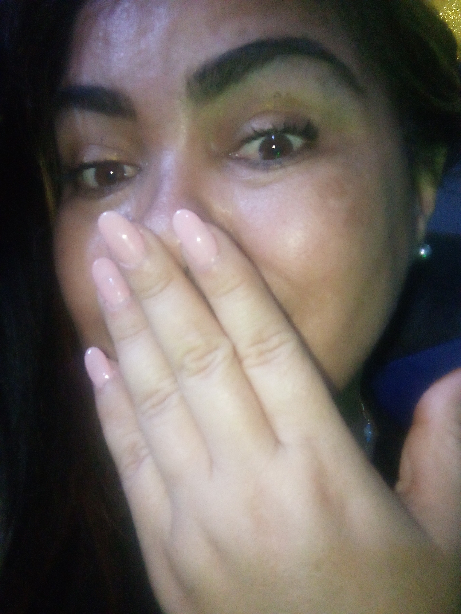 A close-up photograph captures a woman partially obscuring her face with her right hand. Her almond-shaped eyes are brown, framed by thick, arched eyebrows and long eyelashes. Her skin tone is tan with apparent blemishes or freckles on her cheek. She has a small pearl earring visible on her earlobe. Her nails are painted a light pink color. The background is dark. There is no information to suggest that this photo was taken in Juan Griego, Venezuela.