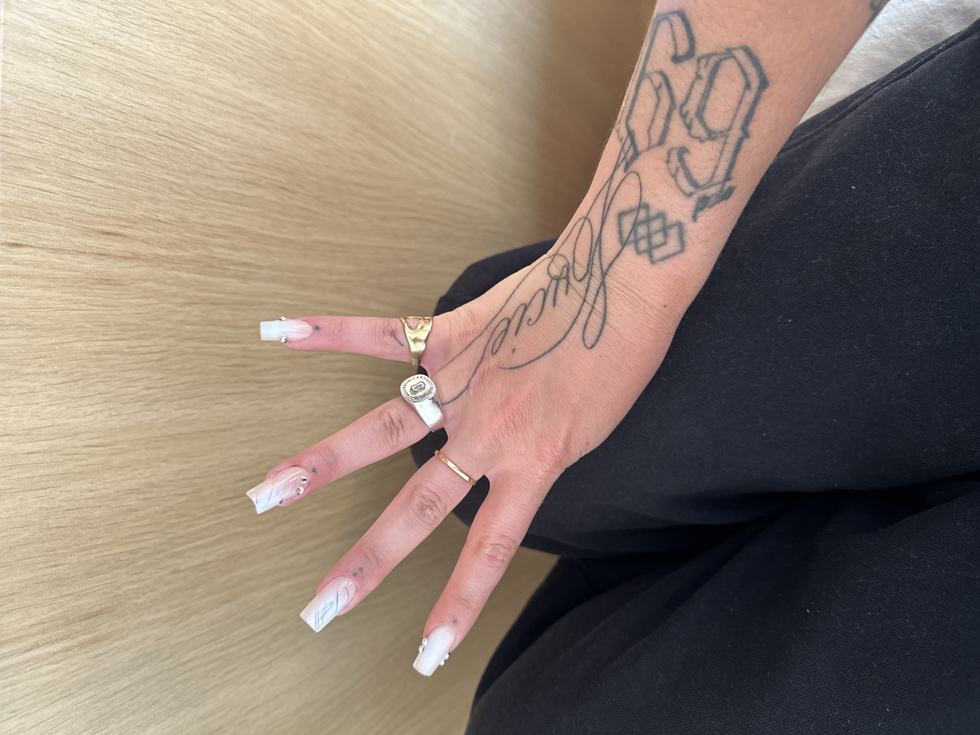 A person's hand is positioned on top of black fabric, likely clothing. The background consists of a light brown, wood-like surface. The hand has several rings: a gold ring on the thumb, a silver ring with a design on the index finger, and a thin gold band on the middle finger. The fingernails are long and squared, with a white design, some adorned with small gems. The person's arm is visible, featuring tattoos. One tattoo reads "Lucie" in script, and another shows the number "69" in a stylized font. The scene does not provide any information about the location in the city of Saint-Ouen-sur-Seine, France.