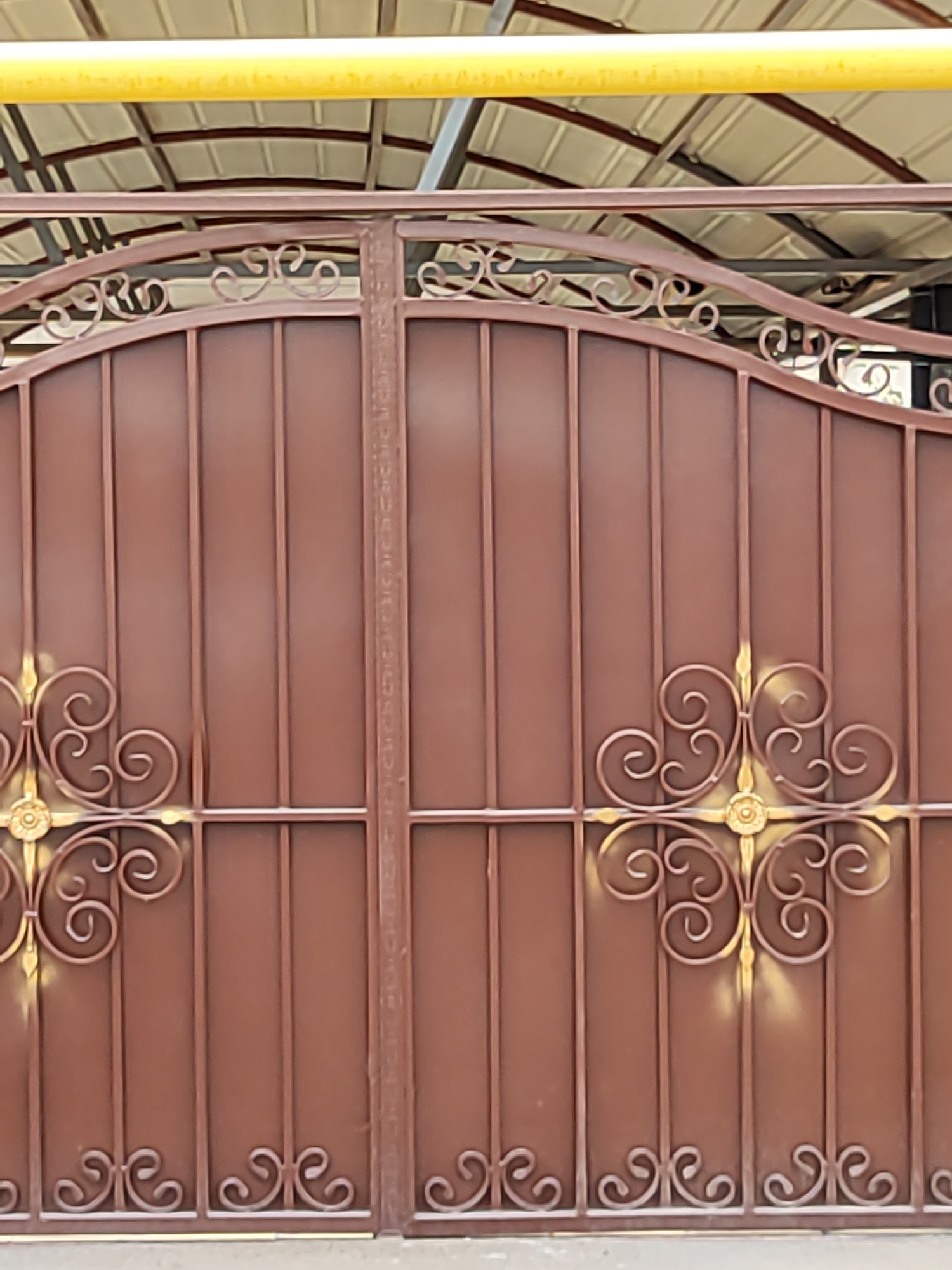 An outdoor medium shot captures a pair of tall, brown, wrought-iron double gates. The gates feature ornate scrollwork, with gold accents highlighting some of the decorative elements. The gates are closed. In the background, a yellow pipe runs horizontally above the gates, and a corrugated metal roof structure is visible. The scene appears to be in the city of Boraldai, Kazakhstan, suggesting a residential or commercial property with an enclosed yard or entrance.