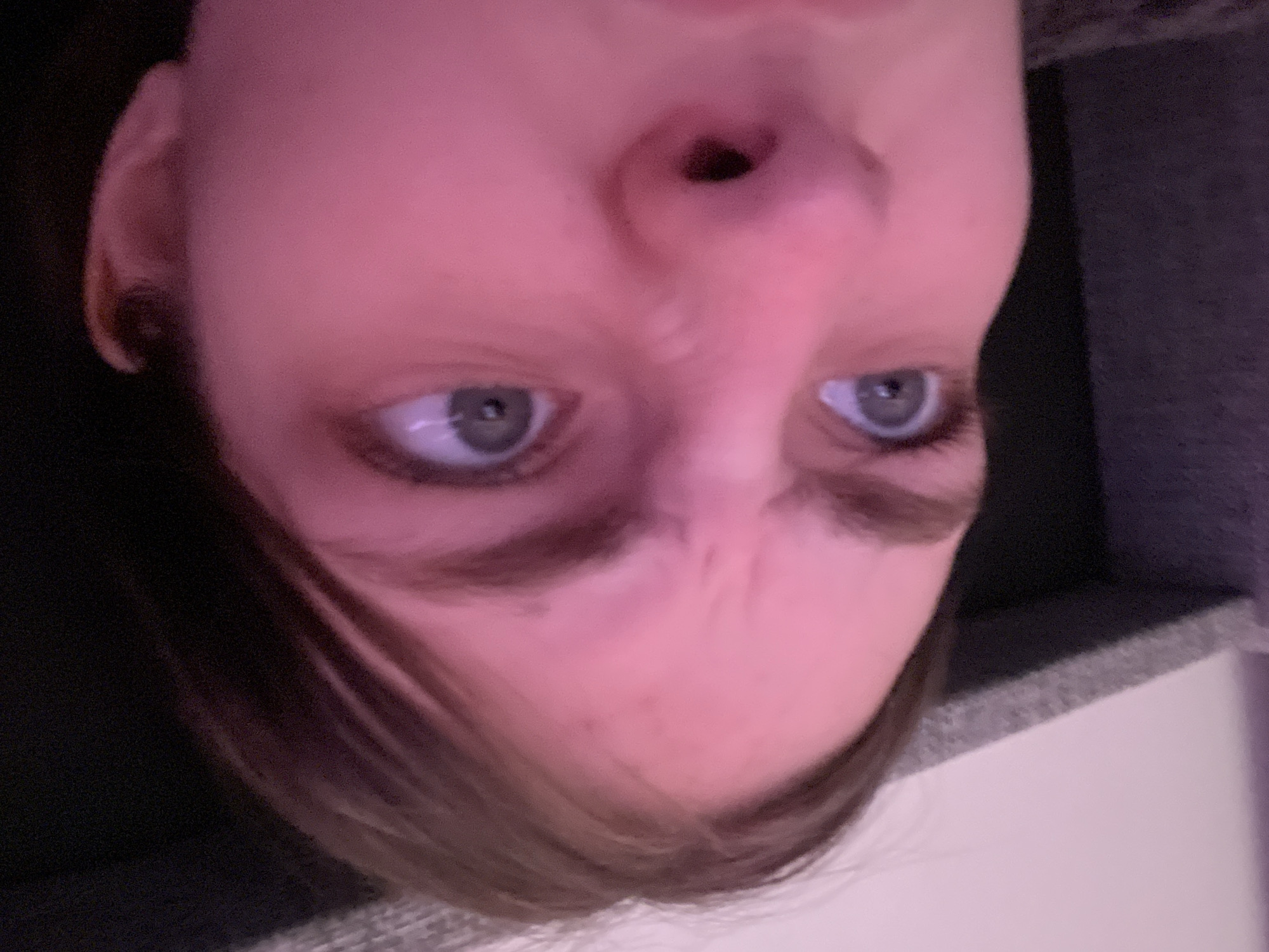 A close-up, slightly out-of-focus image of a person's face, taken from a low angle. The person has light skin, light-colored eyes, and light brown hair. The image is rotated, with the person's face appearing upside down. The person is wearing a dark-colored shirt or sweater. Part of a light-colored object (possibly a wall or furniture) is visible in the lower right corner. No information allows for the determination of a location in Saint Petersburg, Russia.