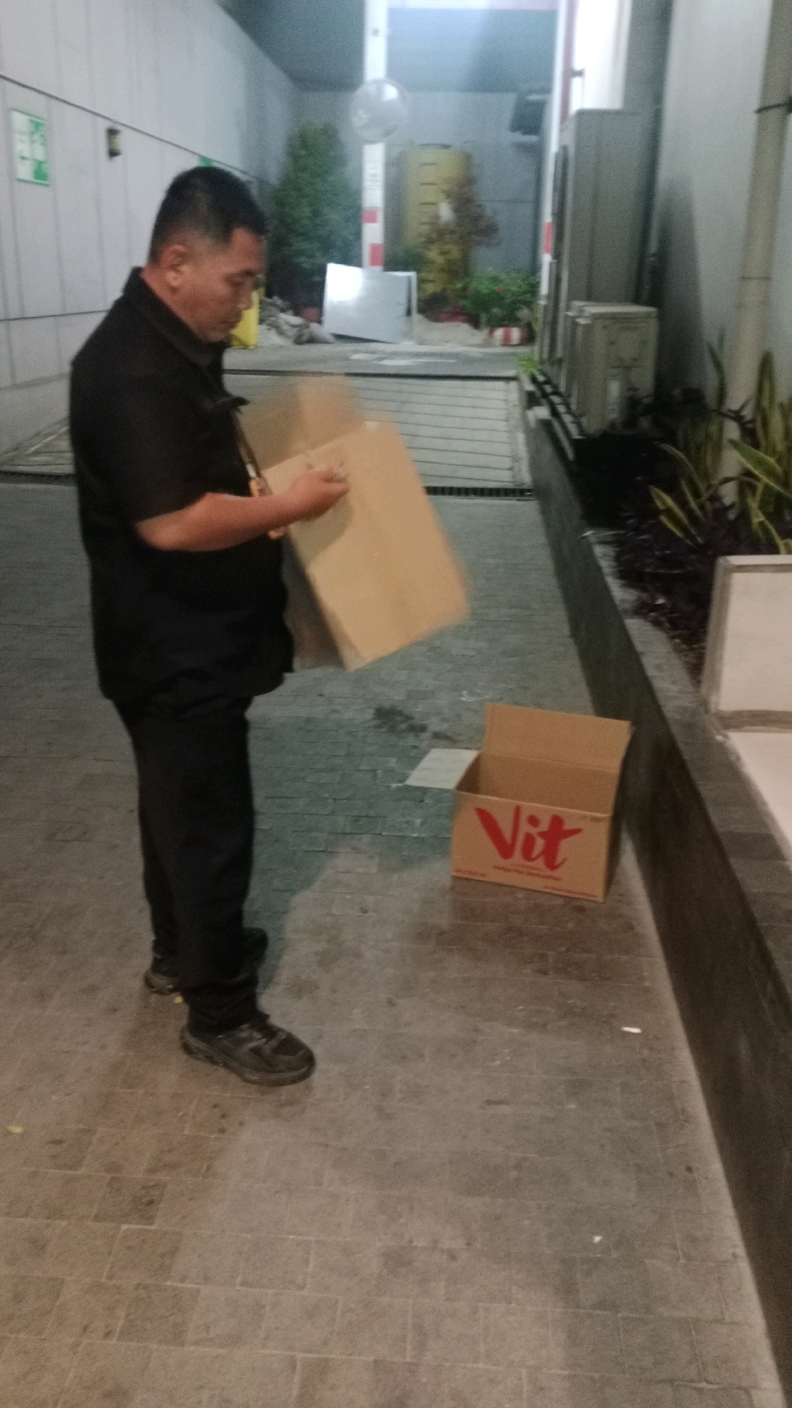 A middle-aged Asian man wearing a black polo shirt and black pants, and black shoes is standing on a tiled pathway. He is holding a cardboard box in his left hand and what appears to be another larger cardboard box in his right hand, held close to his chest, with his right arm bent. The man is facing slightly to his left, looking down at the box he is holding. To his right, on the ground, is an open, smaller cardboard box with the word "Vit" printed in red on its side. The pathway is paved with square grey tiles and shows some damp patches. In the background, there is a wall on the left, and what looks like the entrance to a building or parking area on the right, with some plants and a yellow cylindrical object. The scene appears to be outdoors, possibly in a service area or alleyway in West Jakarta, Indonesia.