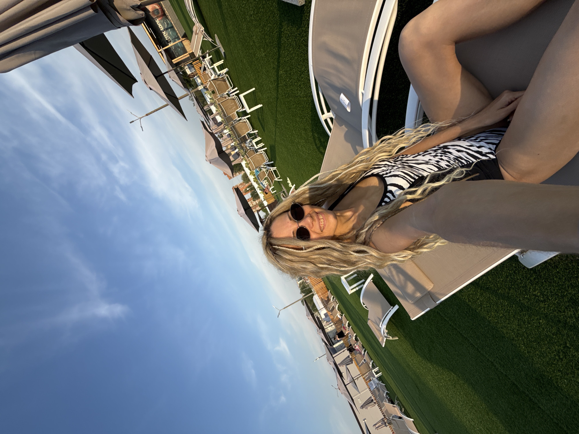 A woman with blonde hair and sunglasses is reclining on a lounge chair in what appears to be an outdoor leisure area. She is wearing a black and white patterned swimsuit. The area has a number of similar lounge chairs with umbrellas, arranged on a green surface that could be grass or artificial turf. The sky is blue with some cloud cover. The location is in Шебунино, Russia.