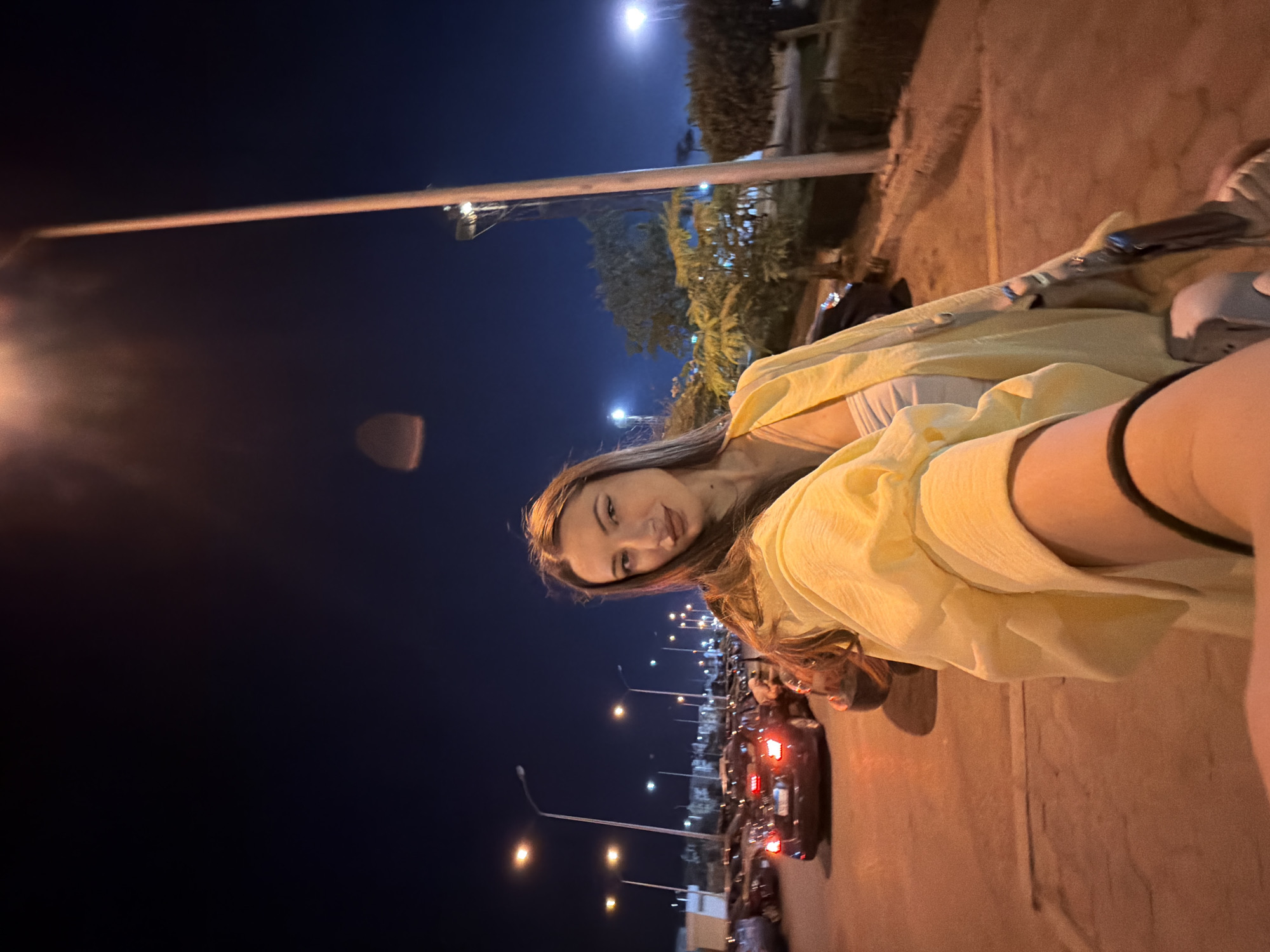 In a nighttime street scene in 6th of October City, Egypt, a young woman with long brown hair and light makeup is taking a selfie. She is wearing a light-colored top and an oversized yellow shirt. The background shows a street with several parked cars, streetlights, and trees. The overall lighting suggests it is evening or nighttime.