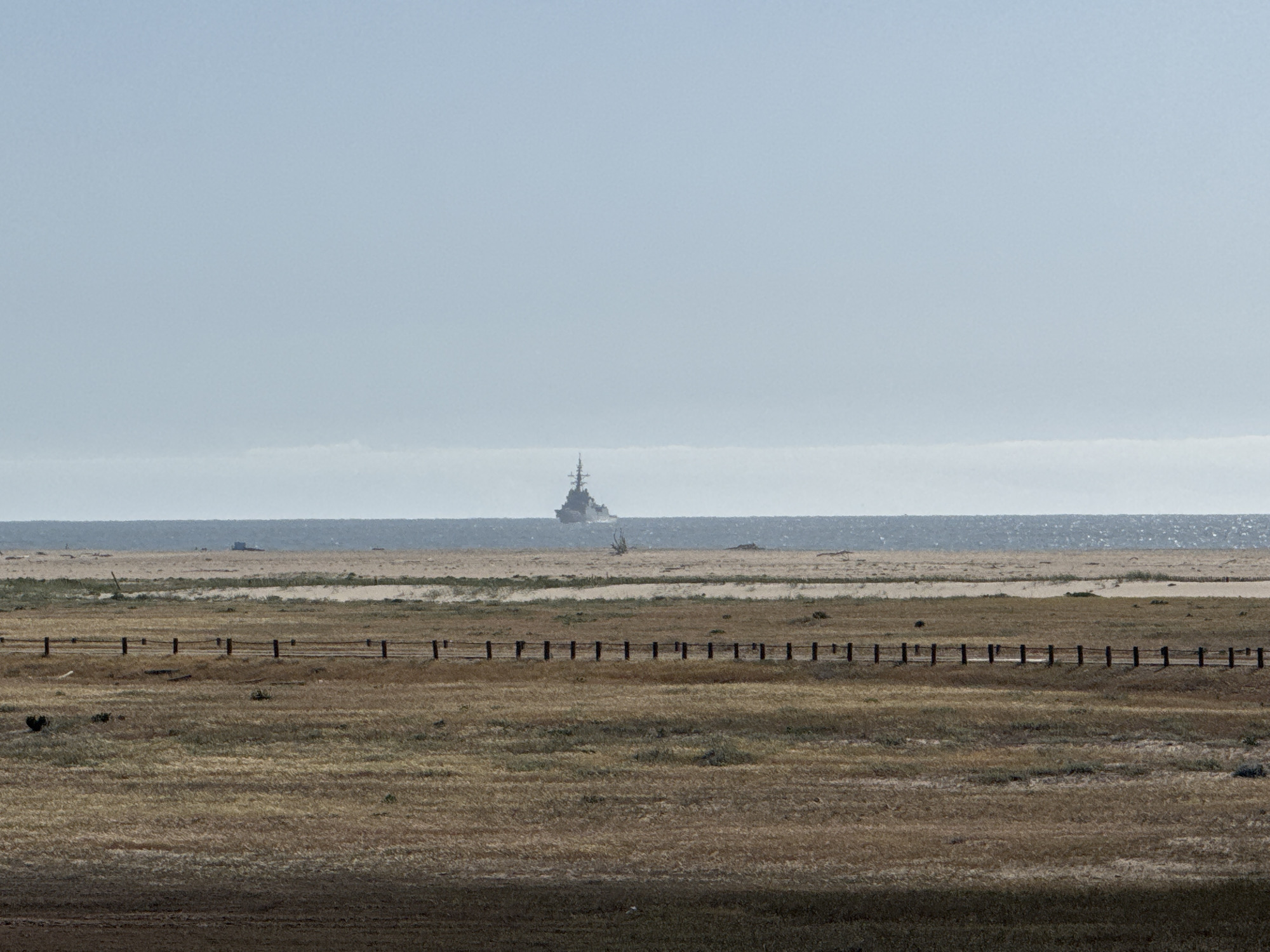 This wide-angle image depicts a coastal landscape in Figueira da Foz, Portugal, under a soft, hazy sky. The scene is dominated by natural elements and a distant vessel.
In the immediate foreground, a broad expanse of dry, golden-brown grass and patchy soil stretches across the lower portion of the frame. This field shows signs of arid conditions, with some darker, bare earth visible.
A simple, rustic fence, constructed from dark wooden posts and a single horizontal chain or wire, runs horizontally across the midground, separating the dry field from a sandy, dune-like area beyond. This sandy strip appears to be a beach or a section of coastal dunes, gently rising towards the water.
Further back, the vast expanse of the ocean, likely the Atlantic, meets the horizon. The water surface shows subtle glints of sunlight, indicating a clear, though perhaps hazy, daytime. Prominently positioned on the horizon line, a large grey naval ship, possibly a destroyer, is visible. Its superstructure, mast, and general profile are clearly discernible against the lighter sky, suggesting it is a significant distance offshore. The ship appears to be underway, possibly moving across the frame from right to left.
The sky above is a uniform, pale blue-grey, contributing to the hazy atmosphere but allowing for clear visibility of the distant ship. There is no discernible specific activity other than the ship's presence at sea. No people or visible text are present in the image. The overall impression is one of peaceful, expansive coastal observation.