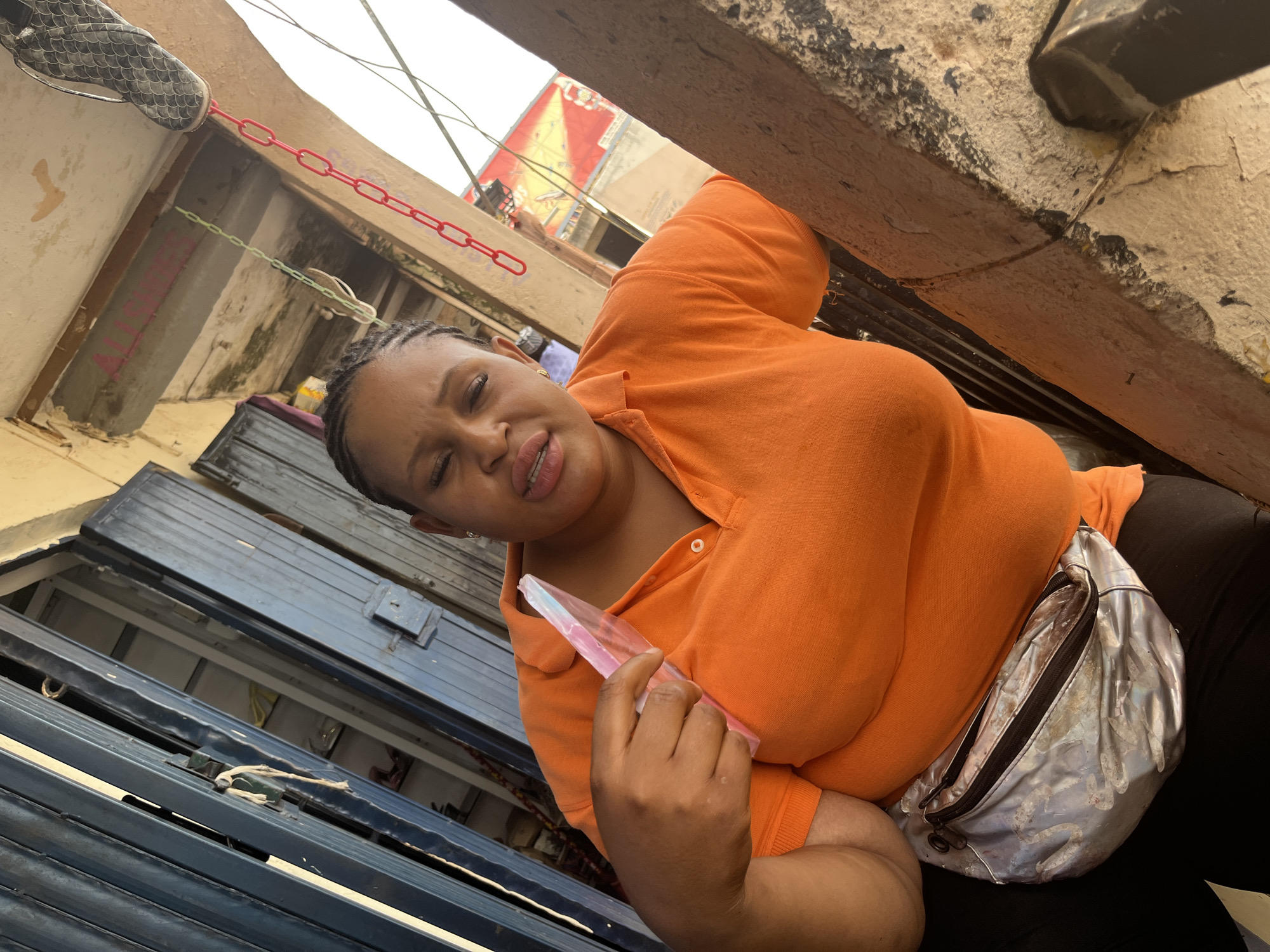 A medium shot shows a Black woman in an orange polo shirt with a silver fanny pack around her waist. She is looking down and holding a pink, plastic object in her right hand. Her braided hair is pulled back, and she is wearing lip gloss. The background appears to be an outdoor market stall in Jos, Nigeria, with a blue metal shuttered doorway on the left. There are chains hanging from a ceiling in the background. The lighting is bright, suggesting daylight.