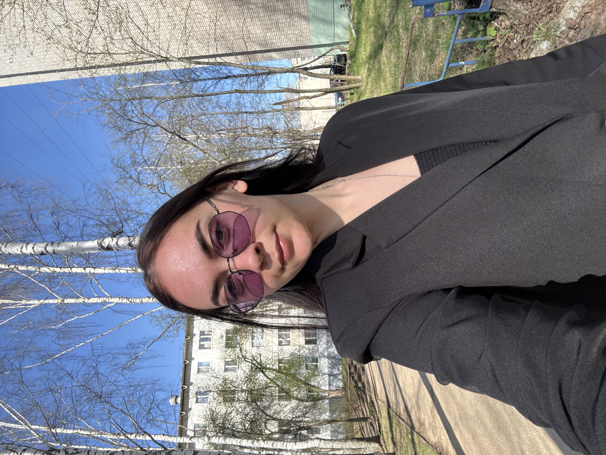 A young woman poses for a selfie outside in the city of Chaikovsky, Russia. She is wearing a black blazer and round sunglasses with purple lenses. Behind her, there's an apartment building, trees, and a clear blue sky. The area appears to be a sidewalk or walking path.