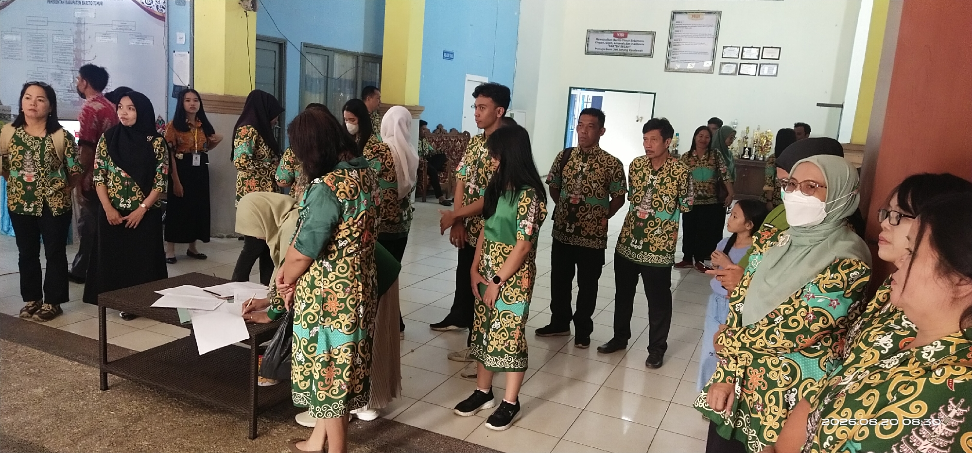 In the city of Tamiang Layang, Indonesia, a group of people, mostly wearing matching batik shirts in green with gold and orange patterns, are gathered indoors, possibly in a government office or community center. The room has a tiled floor and light-colored walls, with some framed posters and charts visible.
In the foreground, a woman with glasses wearing a light green headscarf and a white face mask is looking to her right, with another woman with glasses standing beside her, partially obscured. Several other individuals, including men and women, are dressed in the distinctive batik shirts and black pants or skirts, standing in small groups. Some are looking towards the camera, while others are engaged in conversation or activity.
To the left, a table with papers and a pen is set up, and a person wearing a light brown headscarf is leaning over it, appearing to write or sign something. On the table, there are stacks of papers, some loose sheets, and a pen. The background shows more people, a doorway, and some trophies or decorative items on shelves. The overall atmosphere appears to be a formal gathering or event. The date stamp "2026.08.20 08:30" is visible in the bottom right corner of the image.