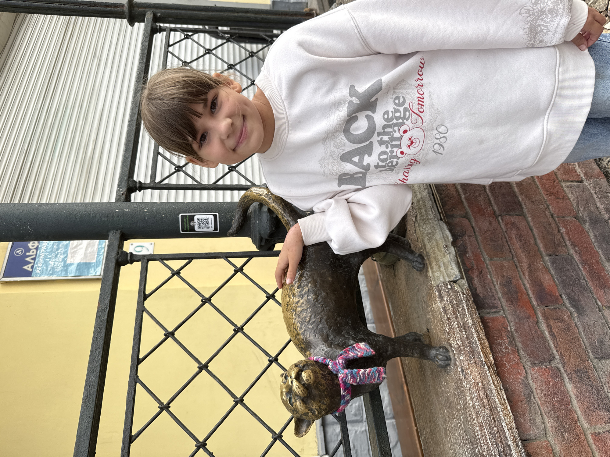 A young girl poses outdoors, smiling brightly directly at the camera. She has light brown hair with bangs and is wearing a white long-sleeved sweatshirt and blue jeans. Her right hand rests gently on the back of a bronze or dark metal cat statue, which features a colorful knitted scarf (pink, blue, and white stripes) tied around its neck.
The setting appears to be an urban or town environment. Behind the girl and the statue, there's a black metal fence with a diamond grid pattern. To the left, a light yellow building wall is visible, on which a blue and white sign with Cyrillic text is mounted. The sign prominently displays "Альфа" (Alfa) and a circled "3," with other smaller Cyrillic script below, consistent with the given location context of Заволжье, Russia. A small QR code sticker is also affixed to a black vertical pole near the fence. The ground or a lower wall surface to the right features distinct red brickwork. In the background, a white corrugated metal surface suggests a building facade or shutter.
The girl's sweatshirt features grey and red text: "BACK to the... (obscured word, possibly 'Future' or 'Forge') chasing tomorrow 1980," along with a cartoon bear head graphic. The time of day appears to be daytime, likely overcast given the soft lighting. The overall mood is cheerful, with the girl's happy expression and casual interaction with the public art.
