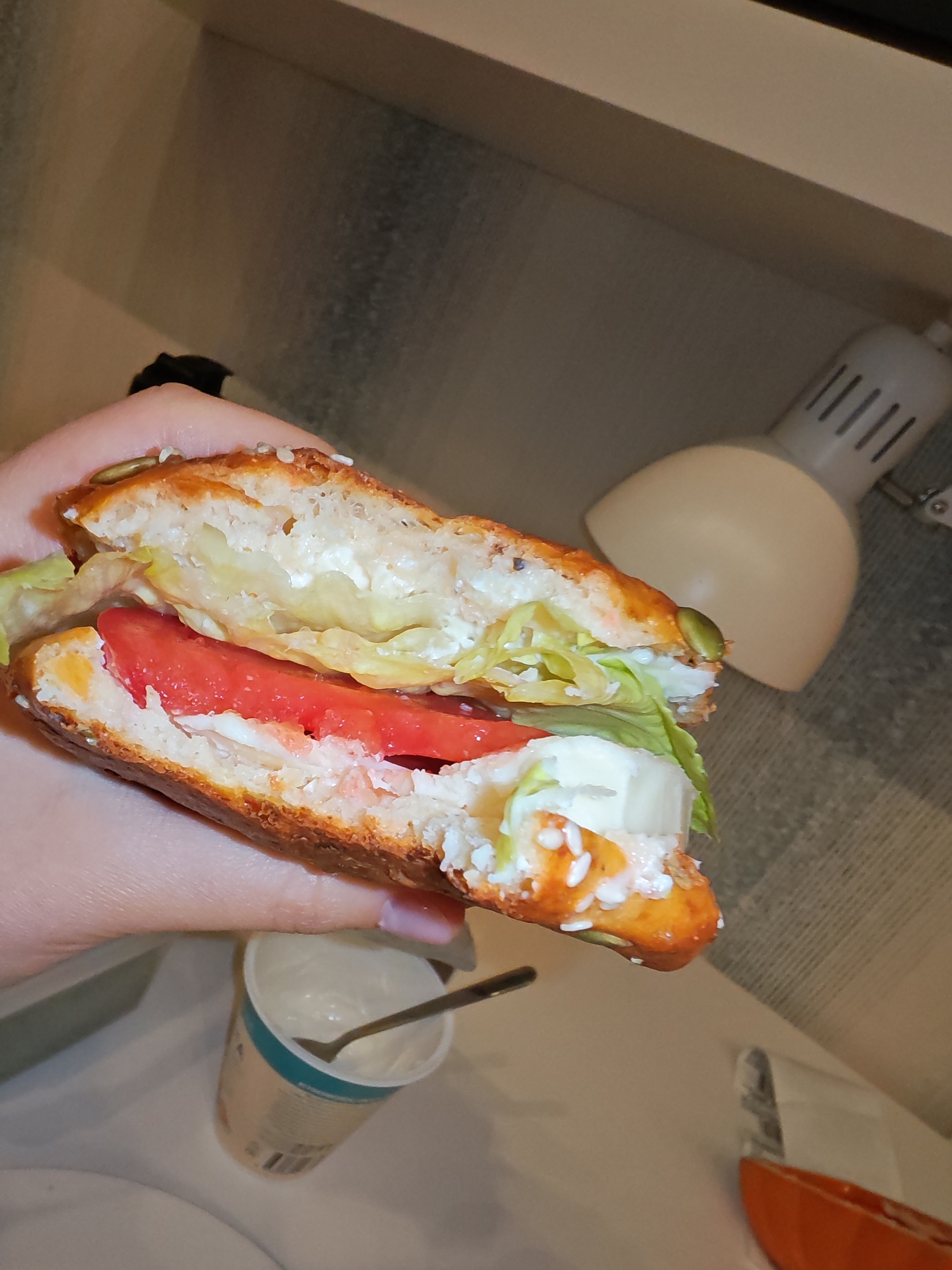 A close-up, eye-level shot shows a person's hand holding a halved sandwich. The sandwich has a light-colored, sesame-seed-covered bun, and the filling includes lettuce, tomato slices, and a creamy white spread. A small metal spoon rests in a white plastic container in the background, and a beige desk lamp is visible behind the sandwich. The scene appears to be indoors, likely in a domestic or office setting. The image is not location-specific to Bataysk, Russia.