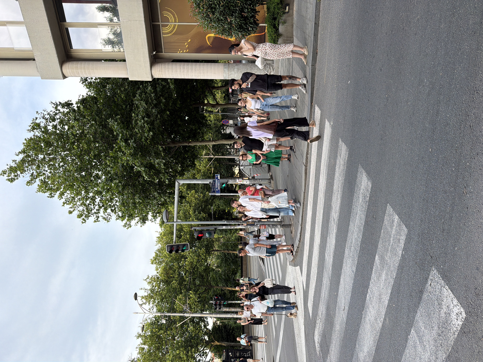 A group of people are gathered at a crosswalk on a street in the Vracar Urban Municipality, Serbia. The sky is overcast, and trees line the sidewalk. A traffic light is visible, showing a red light for vehicles and a green light for pedestrians. Some individuals are waiting to cross, while others are already on the crosswalk. A couple is walking a small dog, and a woman is pushing a stroller. The buildings in the background are modern in style.