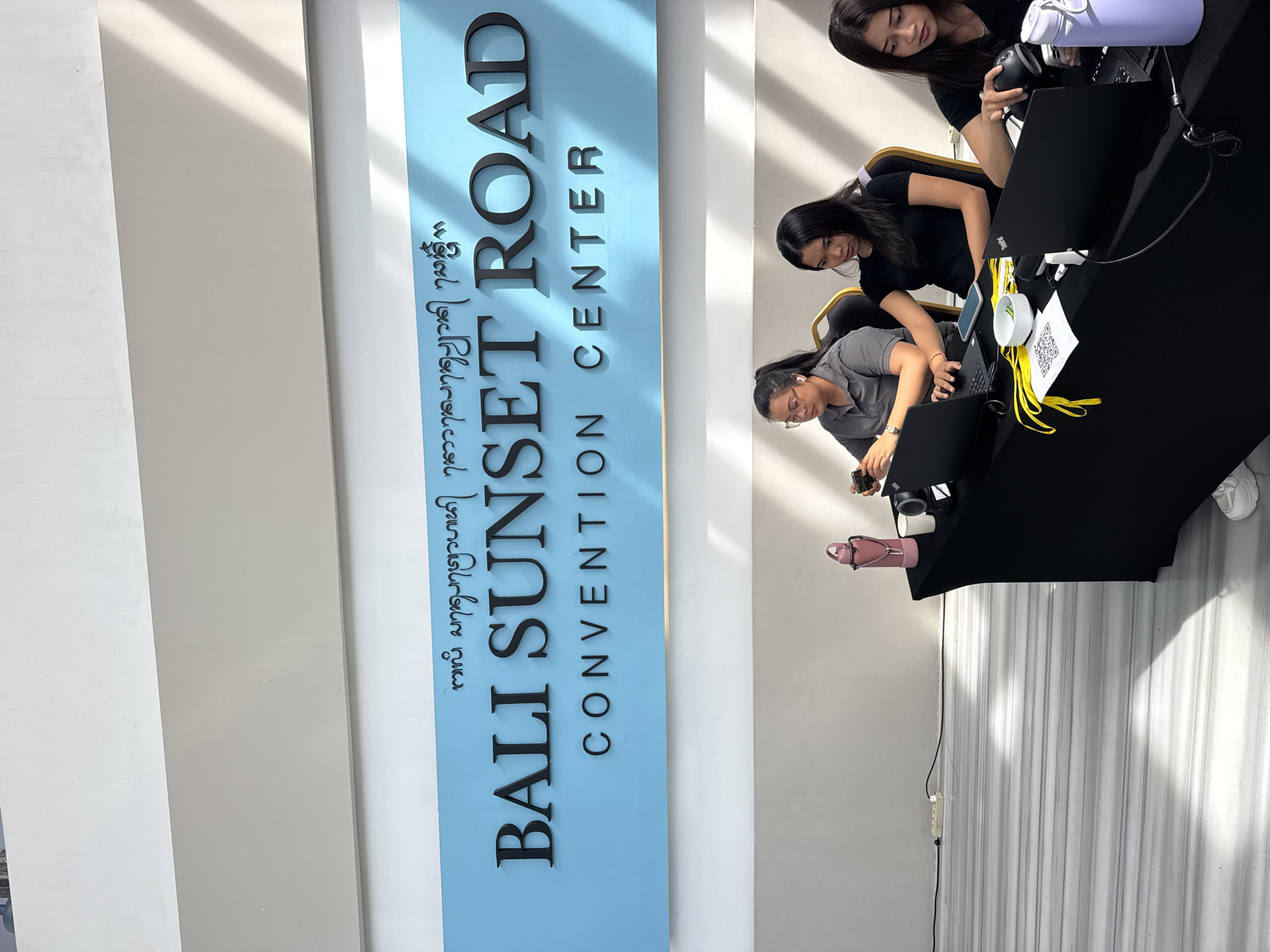 Three women are seated at a table covered with a black cloth, working on laptops and using a mobile phone. The environment appears to be inside the Bali Sunset Road Convention Center in Denpasar, Indonesia, as indicated by the prominent sign in the background. The sign, predominantly light blue with black lettering, reads "BALI SUNSET ROAD CONVENTION CENTER" along with some writing in the local language. Objects on the table include laptops, phones, yellow ribbon, and a QR code tag.