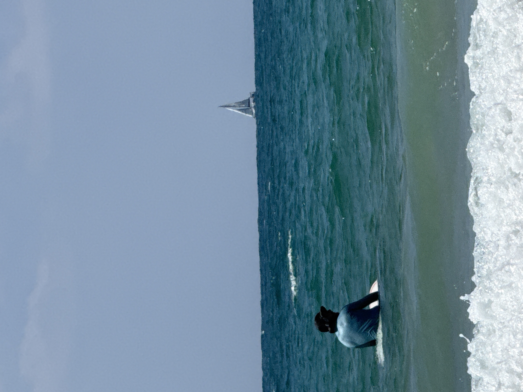 A single person wearing a dark hat and a blue shirt sits on a surfboard in the ocean, facing away from the camera. They are positioned in the lower left quadrant of the image. The water is a deep turquoise color with small waves. In the background, a white sailboat with its sail up is visible on the horizon, sailing on the blue sea under a clear sky with a single wispy cloud. The scene depicts a calm day at sea with a solitary surfer and a distant sailboat. This image is a photograph taken at Gordon Beach in Tel Aviv, Israel, known for its surfing conditions and vibrant atmosphere.