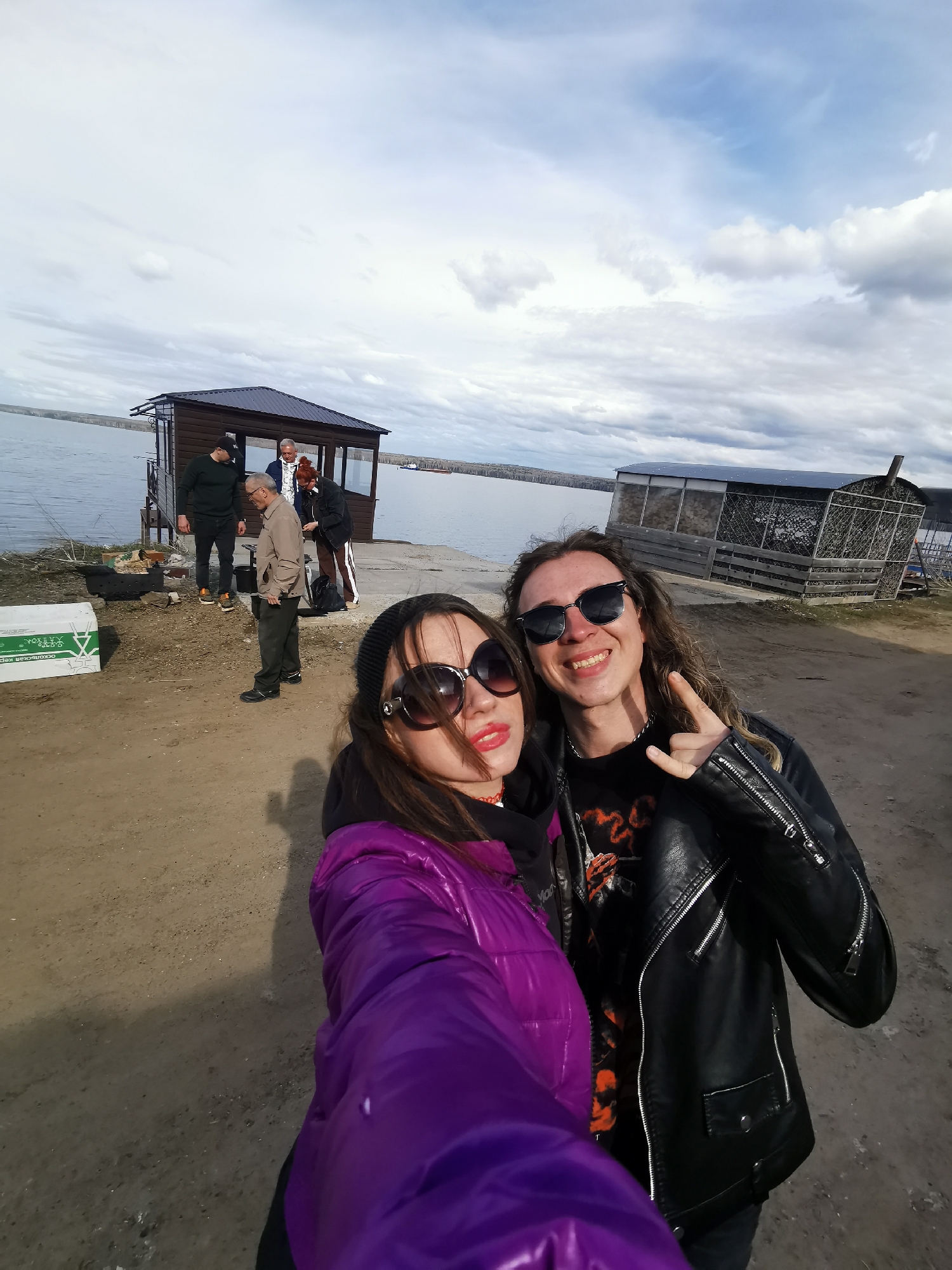 The image captures a casual outdoor scene by a wide body of water under a cloudy sky. In the immediate foreground, two young individuals are taking a selfie, both wearing sunglasses and appearing happy. The person on the left, a woman with dark hair, wears a vibrant purple puffer jacket and has red lipstick visible. She holds the camera for the selfie. Beside her, a man with long wavy hair, donning a black leather jacket over a dark t-shirt with an orange graphic, smiles broadly and makes a "rock on" hand gesture.
In the midground, several other people are gathered on a dirt and gravel patch near a small wooden gazebo or shed with a dark roof, which is positioned at the water's edge. This group includes a man in a dark green top, an older man in a light brown jacket, another man in a lighter-colored jacket, and a woman in a dark jacket with striped pants. They appear to be engaged in a relaxed social gathering, with a visible barbecue grill or fire pit and a white box nearby. The white box has Cyrillic text, including "ОСКОЛЬСКАЯ КРЕПЬ".
The backdrop consists of a large body of water, likely a river or lake, with a distant forested or natural landmass marking the horizon. The sky is overcast with numerous clouds, indicating daytime conditions but diffused sunlight. To the right, further along the shoreline, there is another rustic, elongated structure constructed from wood and mesh. A small vessel, possibly a barge, is faintly visible on the water in the distance. The overall setting suggests an informal recreational area by the water.