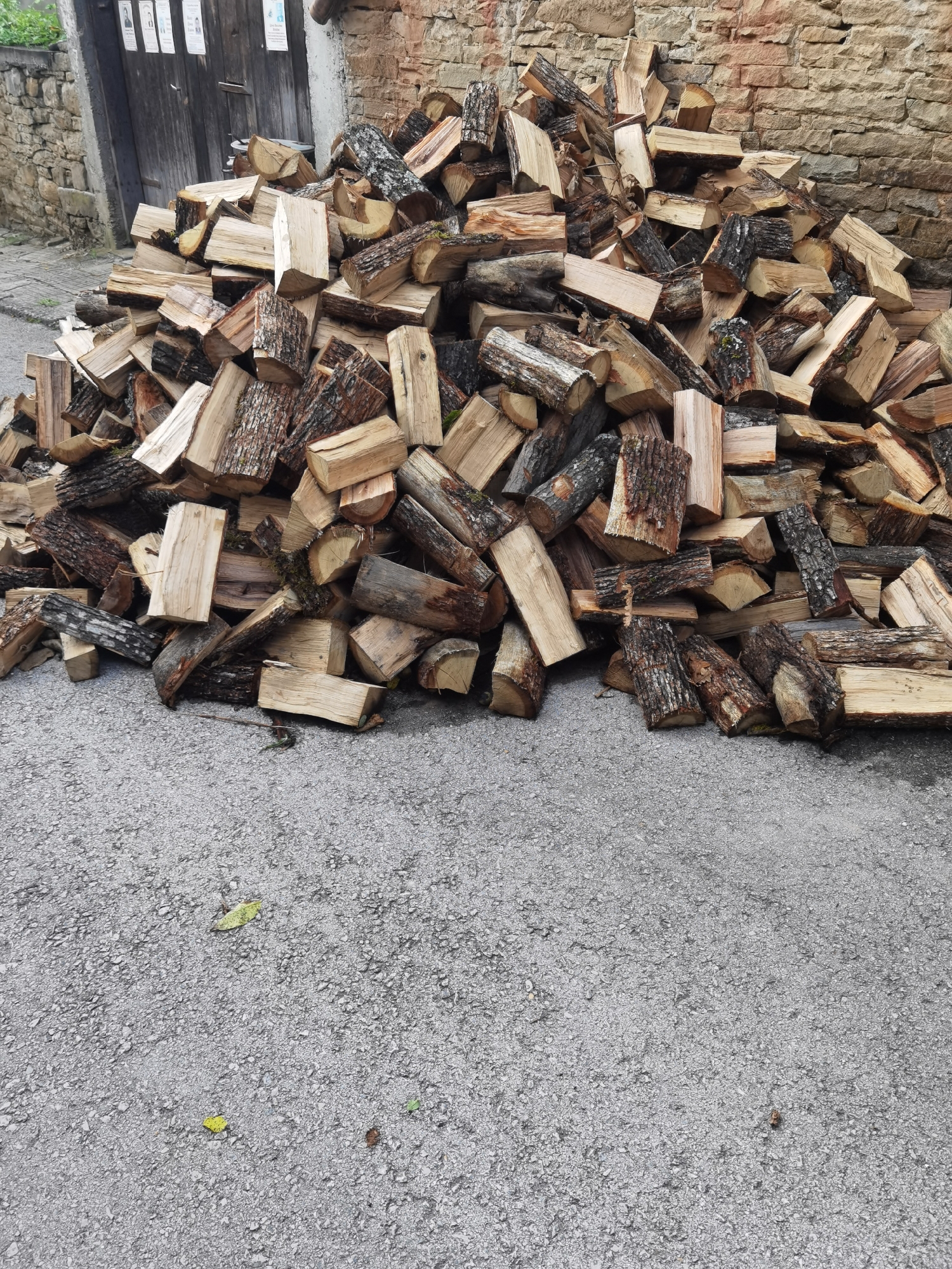 A large pile of chopped firewood is stacked on a grey asphalt surface. The wood is of various sizes and types, with some pieces showing rough bark and others with smooth, cut ends. In the background, a rough stone wall made of beige bricks is visible, and behind that, a weathered wooden door. The environment appears to be outdoors, likely in a rural or semi-rural setting, consistent with the village of Lomets, Bulgaria. There are no people or animals in the image. The lighting suggests it is daytime, with some shadows cast by the wood.