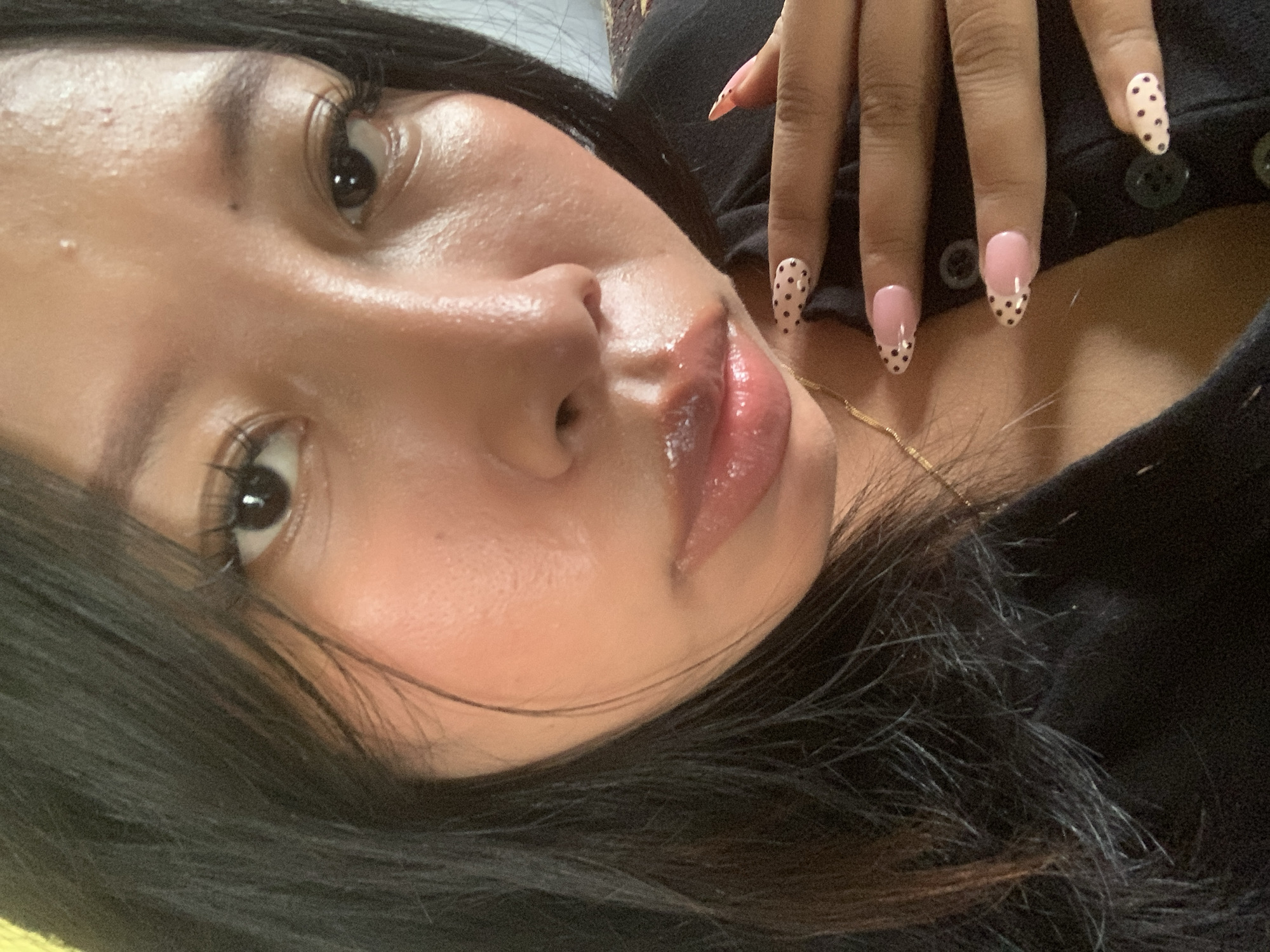 A close-up, selfie-style shot shows a young woman with dark hair, a fair complexion, and dark eyes. She is wearing a black top and has long, sculpted fingernails with a pink and black polka dot design. The woman's face is slightly turned, and her lips are glossy. She is resting her hand on her chest, and a gold necklace is visible. The background appears to be a neutral-toned fabric. The overall impression is of a casual, self-portait moment.