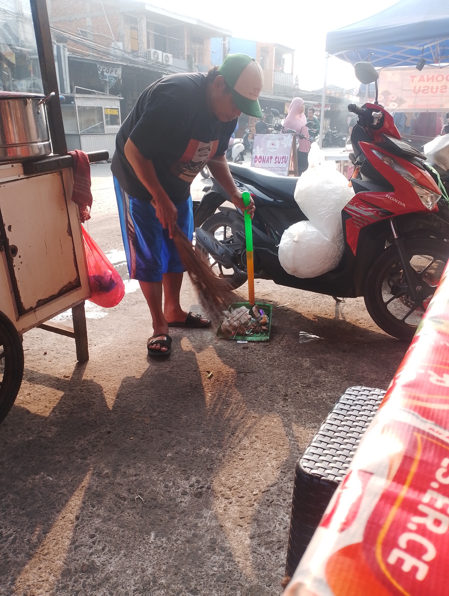In Bekasi, Indonesia, a person wearing a black t-shirt, blue shorts, and a green baseball cap is sweeping the ground with a broom and dustpan. The person is standing next to a food cart with a metal pot on top and a red bag hanging from it. A red Honda motorcycle with two large white bags on the seat is parked nearby. In the background, there are buildings, a blue tent, and other people. The scene appears to be a street-side market or food stall area.