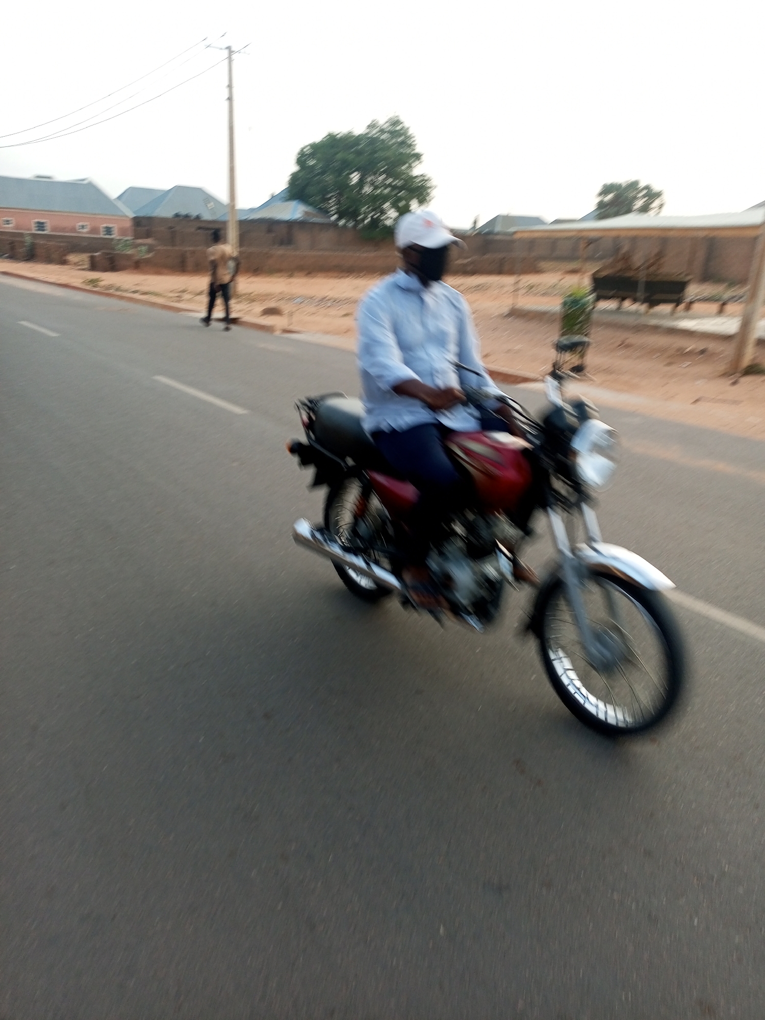 A person wearing a white cap and a black face mask is riding a red and black motorcycle on an asphalt road. In the background, there are houses, trees, and a person walking on the side of the road. The location appears to be a street in Tempure, Nigeria.