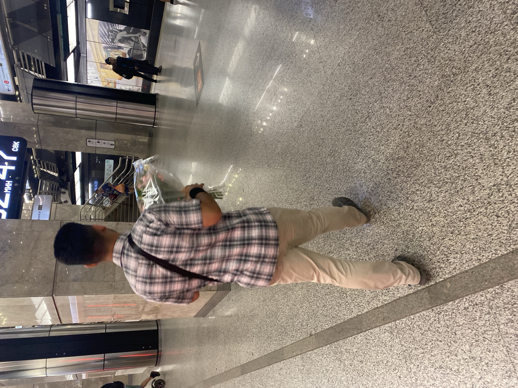 A person walks away from the camera, holding a bouquet of flowers wrapped in plastic. They are wearing a plaid short-sleeved shirt, khaki pants, and dark shoes. The person has a bag slung across their body. The background is an indoor space with polished floors, large pillars, and signage indicating a transportation hub, possibly a subway station. Other people and escalators are visible in the background. The lighting is artificial and overhead.