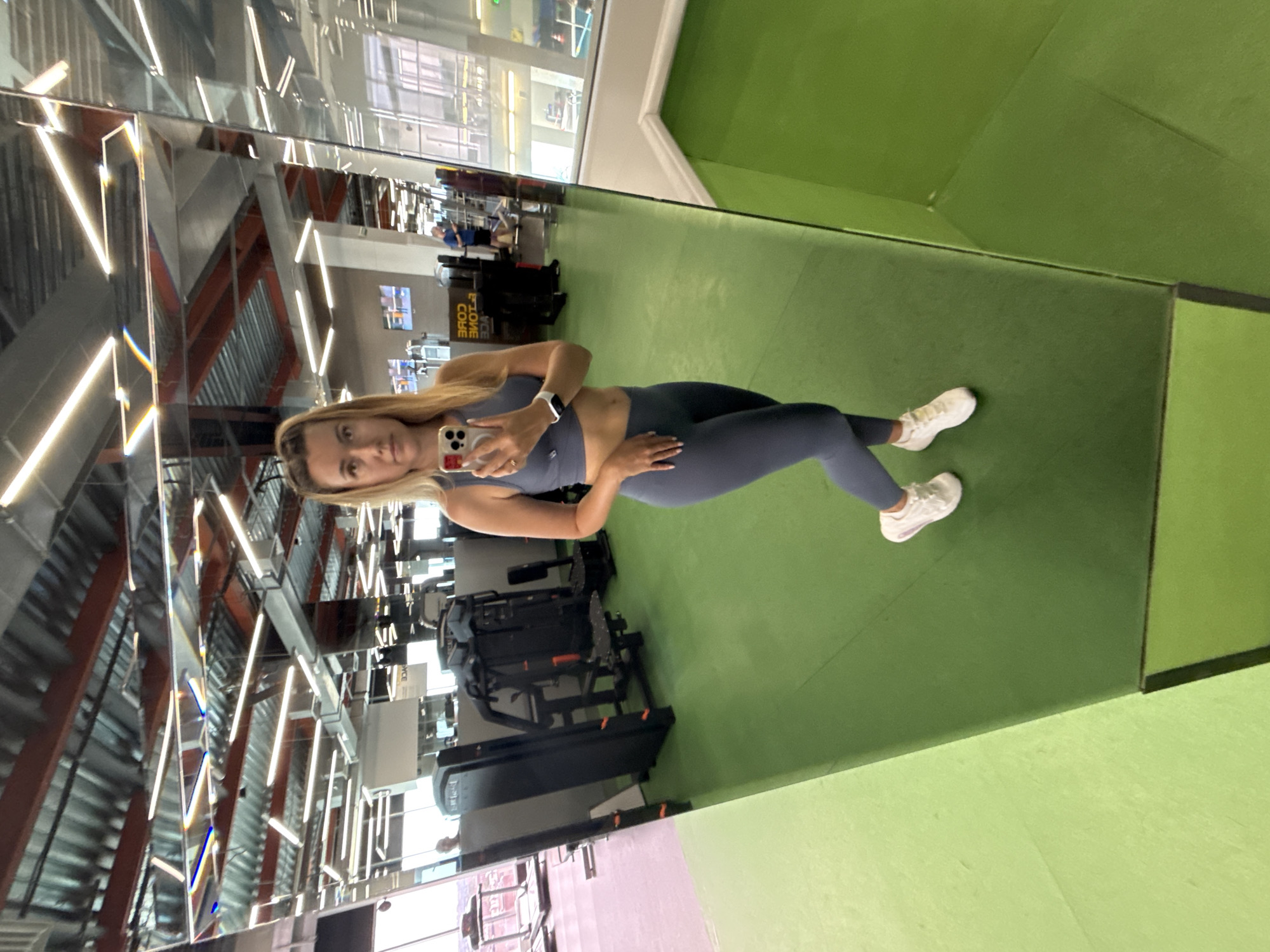 A woman is taking a mirror selfie in a gym. She is wearing a grey athletic set consisting of a sports bra and leggings, and white sneakers. Her hair is blonde and pulled back. She is holding a smartphone in her right hand, and her left hand is resting on her hip. The gym has green flooring and various exercise machines are visible in the background, along with mirrors on the ceiling and walls. The lighting in the gym is bright, with fluorescent lights.