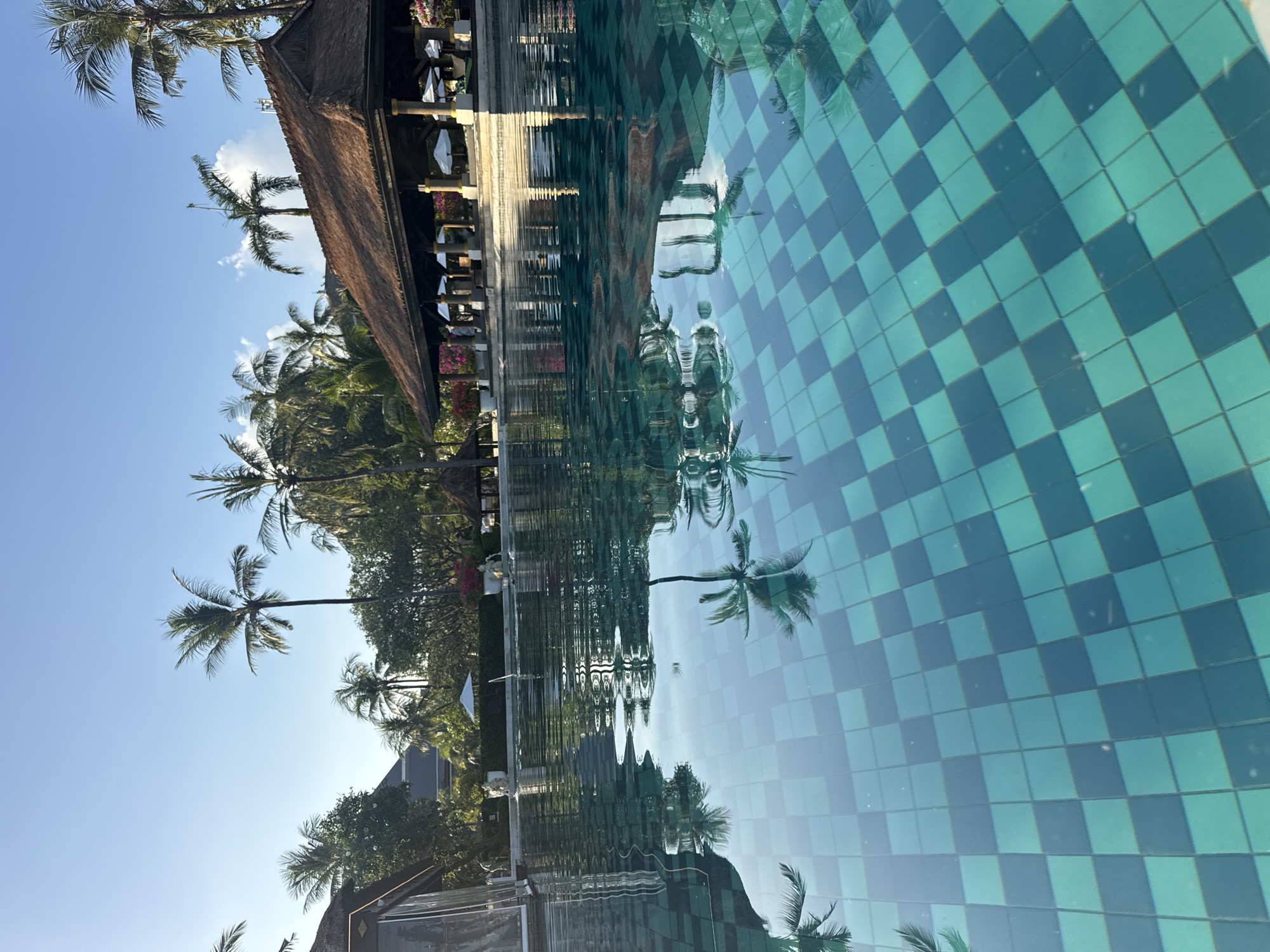 The image shows a pool in Kuta Selatan, Indonesia with a checkered tile pattern on the bottom. Reflections of palm trees, a building with a thatched roof, and the sky are visible in the water. The sky is blue with a few clouds.