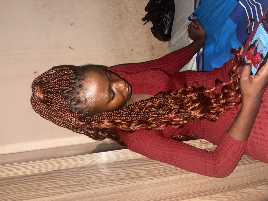 A woman in a red long-sleeve dress is lying down, possibly on the floor, next to a bed with blue and white striped sheets. Her head rests on the floor and she is holding a cell phone, looking at something on the screen. She has copper-colored braided hair with curly ends. A black plastic bag is visible on the bed behind her. The lighting is somewhat dim.
