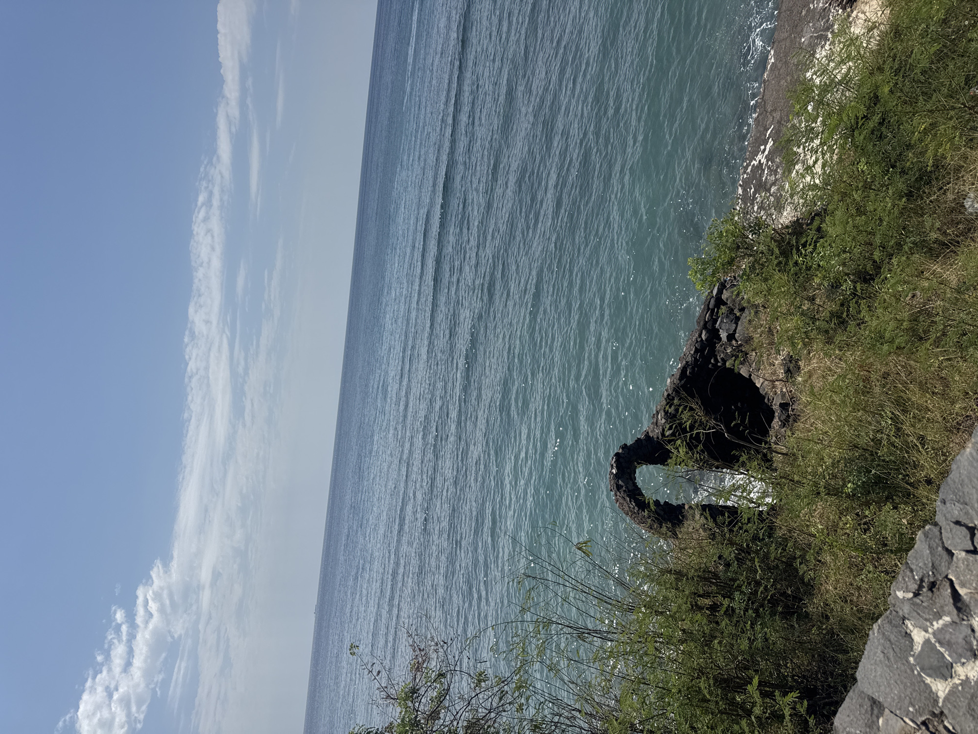An arched stone ruin, partially overgrown with vegetation, stands on a rocky outcrop overlooking a bright blue ocean under a clear sky with wispy clouds. The water exhibits gentle ripples and reflects the sunlight. In the distance, a faint ship is visible on the horizon. The foreground features a stone wall constructed from dark, angular rocks. The scene is located on a cliffside, suggesting a coastal environment. The vegetation consists of grasses and leafy bushes, with some bare branches extending from the left side of the frame. There are no people or animals present in the image. The overall impression is one of natural beauty and historical remnants. This location is in the city of Abang, Indonesia.