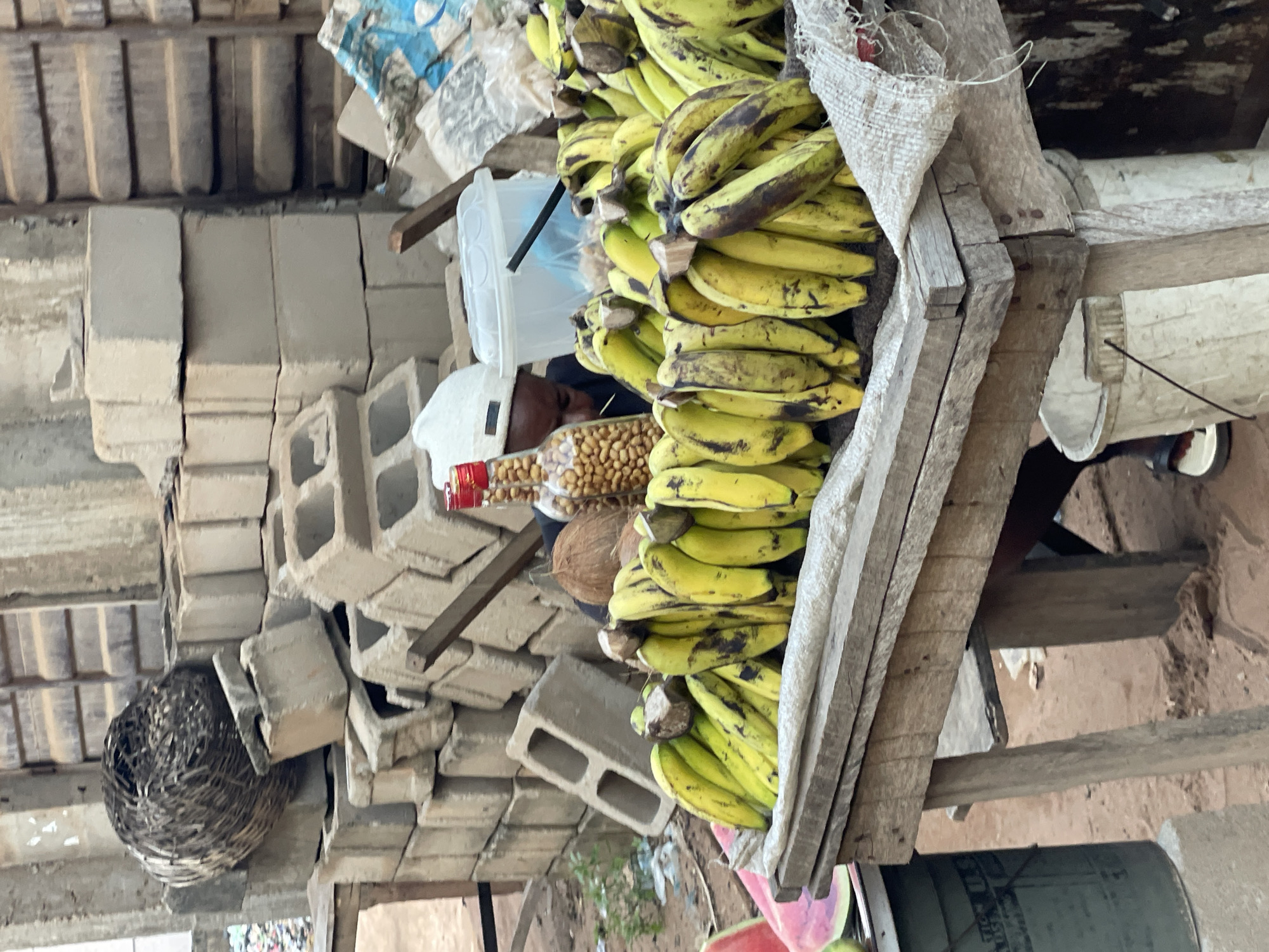 The image depicts a roadside market scene in Nkpor, Nigeria. A large pile of ripening plantains sits on a wooden stall. Behind the stall are stacks of concrete blocks and a woven basket. A vendor wearing a white cap is visible behind a clear plastic container and a jar of nuts, suggesting these are also for sale. A coconut also sits on the display. The stall appears to be makeshift, constructed from rough-hewn lumber and a burlap sack.