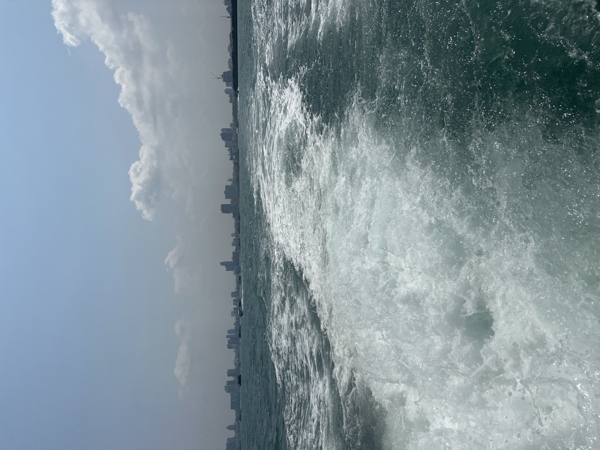 The image shows the sea water of Pattaya City, Thailand, with a city skyline visible in the background. The sky is partially cloudy, with a pale blue hue. The sea is choppy, with visible waves and foam, suggesting movement, possibly from a boat.