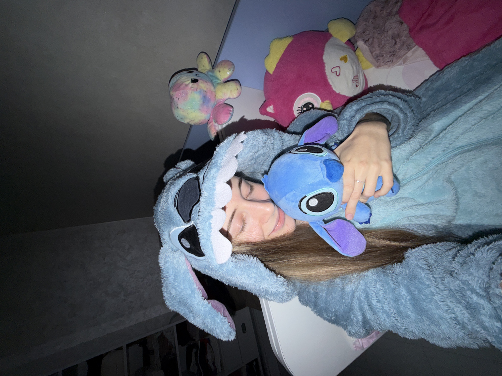 A young woman is lying down, wearing a light blue "Stitch" onesie. She is cuddling a blue "Stitch" plush toy. Behind her are several other stuffed animals: a pink animal with yellow details, and a colorful octopus-like toy. The setting appears to be indoors, possibly a bedroom. There is no evidence that the location is in Plovdiv, Bulgaria.