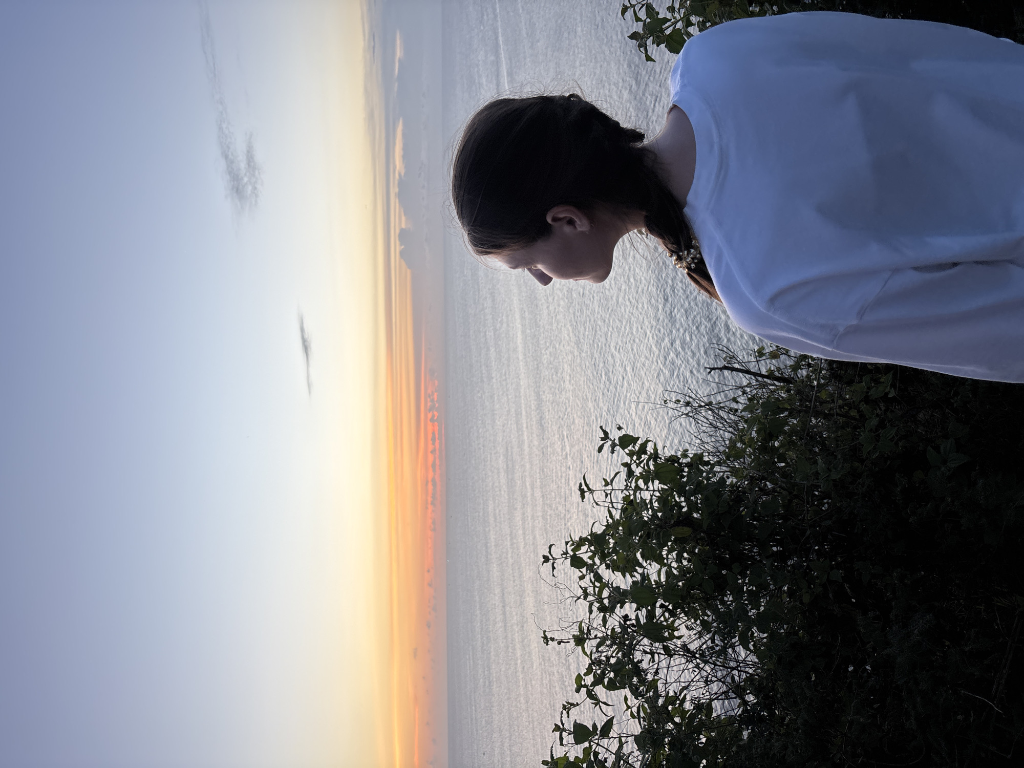 The image captures a person standing on a cliff overlooking the ocean at sunset. The individual, seen from the back and side profile, has brown hair styled in a braid. They are wearing a white, long-sleeved shirt. The background shows a coastal scene with the ocean reflecting the warm colors of the sunset. Vegetation grows along the edge of the cliff. The location is most likely Pecatu, Indonesia, based on the presence of the cliffside vegetation and ocean views.
