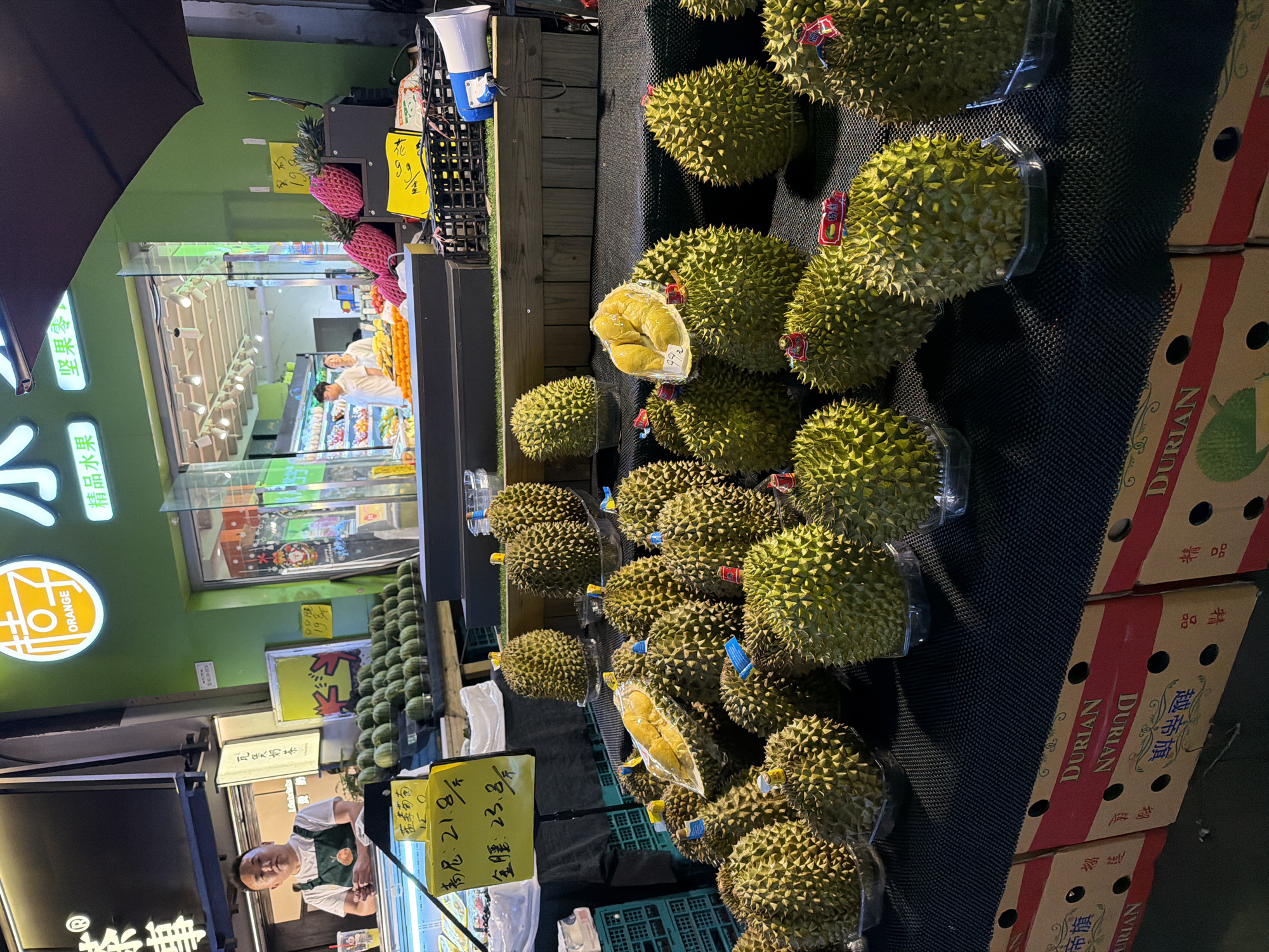 Here is a detailed description of the image:
**Scene:** The image shows a well-lit outdoor fruit market stand, likely at night. The location appears to be in the Chongqing High-tech Zone, China, based on the Chinese text on signage and packaging.
**Subjects/Objects:**
* **Durians:** The primary focus is on the large display of durians, some whole and some cut open to reveal the yellow fruit inside. They are positioned atop a black mesh surface with boxes of durians stacked nearby.
* **Other Fruits:** There are also pineapples (pink) and other round green fruit (likely melons) visible in the background.
* **Signage:** Several yellow signs display prices and names of the produce in Chinese. Boxes with "DURIAN" written on them are visible.
* **Vendor:** A man wearing an apron, presumably the vendor, stands behind the display near a sign.
* **Storefront:** The stand is located in front of a store with a window displaying fruits inside. The store's logo features the word "ORANGE."
* **Other:** There is an umbrella, speakers and a cashier desk.
**Actions/Interactions:**
* The vendor is presumably attending to the display, ready to sell fruit to customers.
* The market is open for business.
**Environment:**
* The environment appears to be a commercial area, likely a street market.
* The scene is well-lit, suggesting it is evening or nighttime.