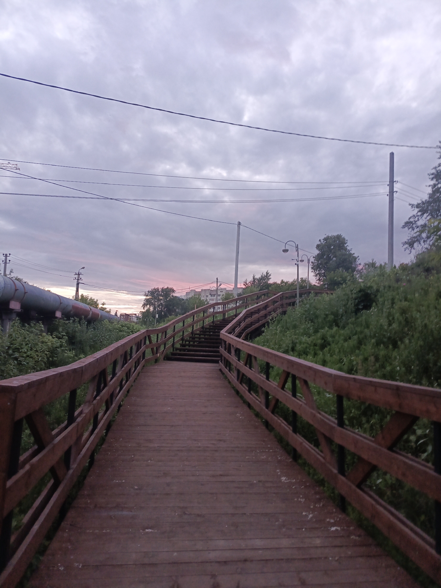 An outdoor, low-angle, vertical shot shows a wooden boardwalk leading up a gentle incline. The boardwalk is made of dark wood planks and is lined with a wooden railing. The railing has a criss-cross pattern formed by diagonal wooden beams connecting to vertical supports.
To the left of the boardwalk, a large, grey industrial pipe runs horizontally, supported by a concrete structure. Behind the pipe, lush green foliage grows densely. Power lines stretch across the sky above the pipe.
To the right of the boardwalk, the incline is covered in thick, green vegetation and trees. Several streetlights with curved arms are visible, spaced along the elevated path. In the distance, buildings can be seen nestled amongst the trees.
The sky above is overcast with a soft, dusky light, tinged with pink and purple hues near the horizon, suggesting either sunrise or sunset. The overall atmosphere is subdued and somewhat industrial, yet softened by the natural greenery and the gentle curve of the boardwalk.
This scene is located in the city of Perm, Russia, specifically in the area known as the "Perm embankment" or "river embankment," a popular recreational area that combines urban infrastructure with natural landscapes. The presence of the industrial pipe suggests its proximity to a thermal power plant or a similar industrial facility that would be found in an industrial zone adjacent to the embankment.