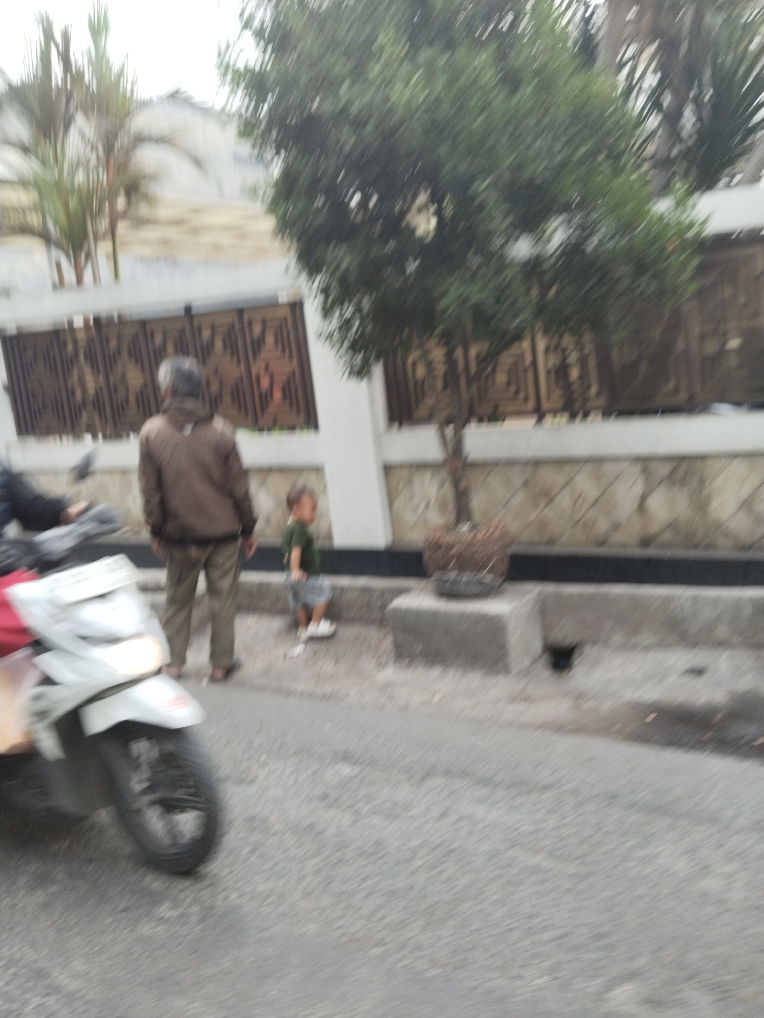 The image depicts an outdoor scene in what appears to be a residential area or along a street. In the foreground, a white motorcycle is visible on the left, partially blurred due to motion. Behind the motorcycle, a person wearing a brown jacket and khaki pants stands facing away from the camera. To the right of this person, a young child wearing a green shirt and a grey skirt or shorts is standing. The child is facing towards the right of the frame. In the background, there is a wall with a decorative pattern and some greenery, including a tree in a pot. The ground is paved with asphalt, and a concrete curb is present. The overall image is somewhat blurry, suggesting it was taken with a moving camera or in low light conditions. Based on the provided metadata, the location is identified as Bandung City, Indonesia.