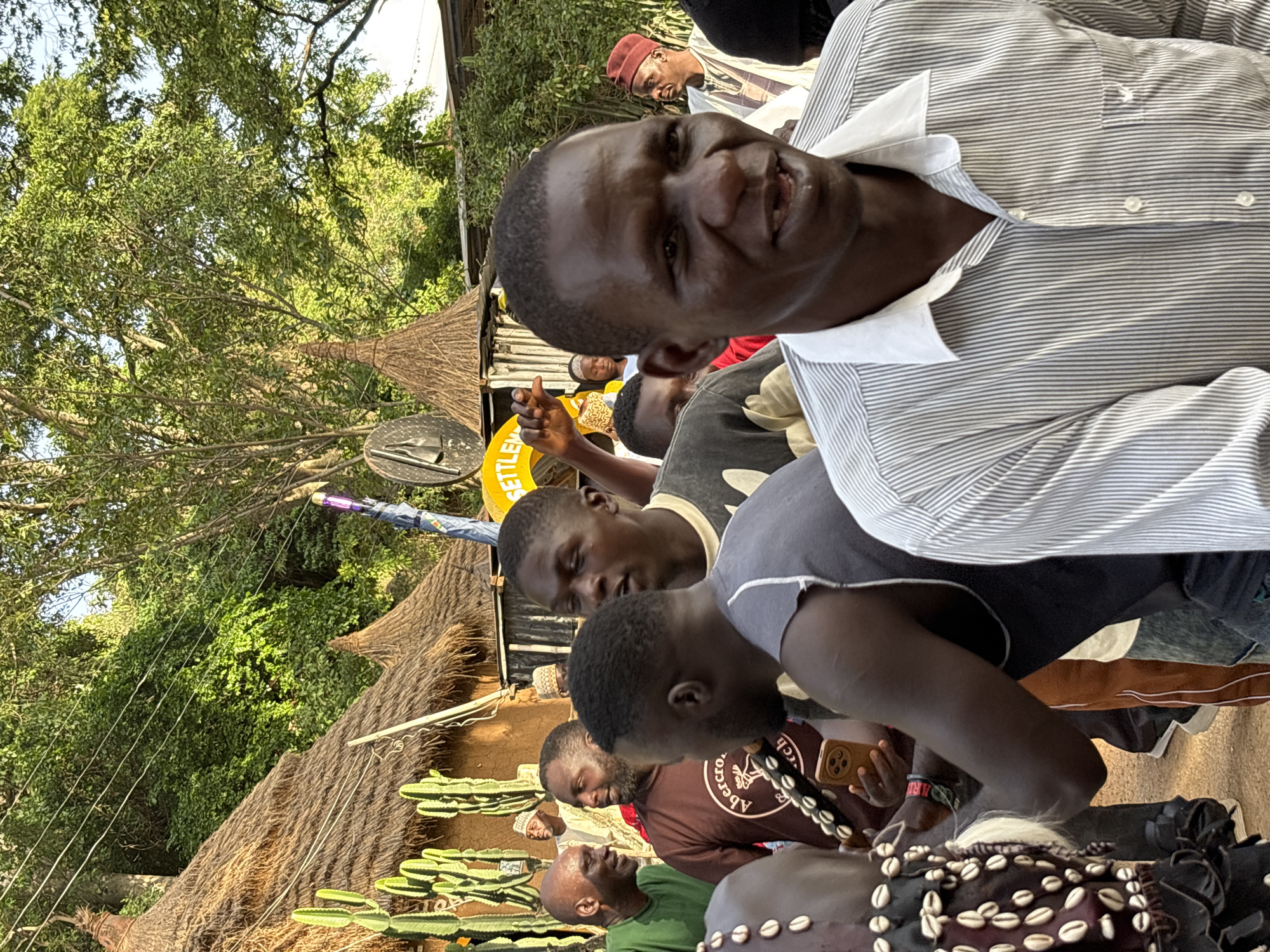 The image captures a gathering of people, primarily men, in what appears to be an outdoor setting. The scene is likely Jos, Nigeria, as indicated. Several men are visible in the foreground, with one man in a striped shirt looking directly at the camera with a slight smile. Behind him, other individuals are engaged in conversation or observing their surroundings.
The setting features thatched-roof structures, possibly huts or market stalls, suggesting a rural or traditional environment. Lush green foliage and trees are abundant, indicating a daytime scene with likely good weather, possibly warm and sunny. Cacti-like plants are also visible, adding to the unique environmental context.
Some of the men are wearing traditional attire, including a man in a red cap and another adorned with a headdress decorated with shells. A yellow circular object with visible text is also present, though the text is partially obscured. A cell phone is visible in one person's hand. The overall mood seems relaxed and communal, with a sense of people coming together.